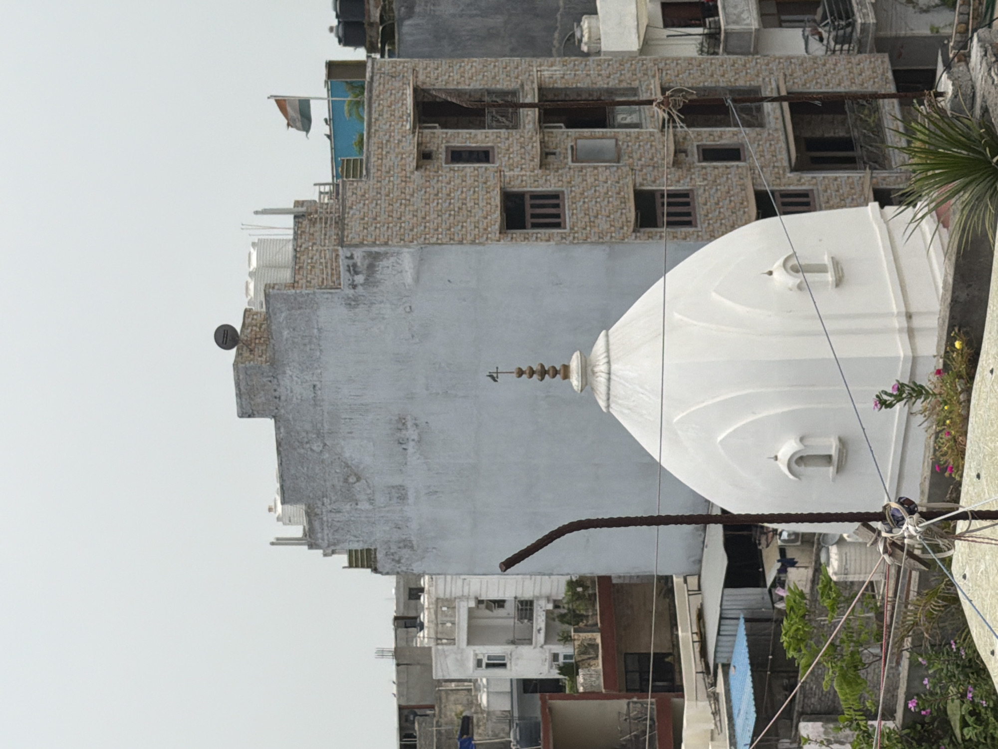 This is an outdoor, eye-level, medium shot of a white domed structure, likely a temple, in a densely populated urban environment. The structure is in the foreground, with a tall, multi-story building with a brick facade directly behind it to the right. The building has numerous windows and balconies. Further in the background, other buildings and rooftops are visible, some with water tanks. An Indian flag is flying from the top of a blue structure on one of the buildings. The sky is overcast and white. The scene appears to be in Delhi, India, as evidenced by the architecture and the Indian flag. There are no people visible in the image. The focus is on the architectural elements and the urban landscape.