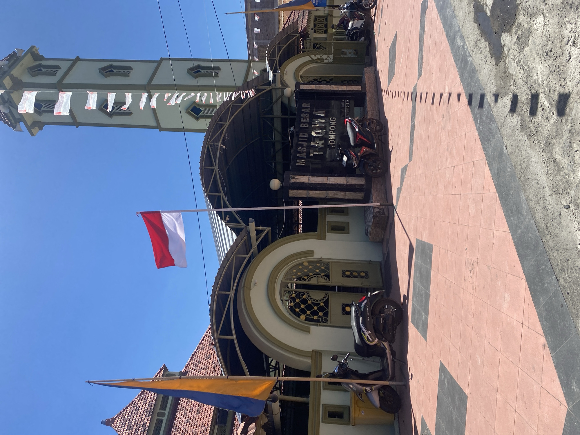 The image shows the exterior of Masjid Besar Taqwa Tompompong, a mosque located in Bantaeng, Indonesia. The mosque features a light green minaret with white decorative flags strung along its height. In front of the mosque, an Indonesian flag, red and white, flies on a pole. Other flags, blue and yellow, are also visible. The mosque has arched entrances and is partially covered by a dark awning supported by a metal structure. Several motorcycles are parked in front of the mosque. The ground is paved with pink and black tiles, with shadows cast by the awning and a pole. The sky is clear and blue, indicating a sunny day.