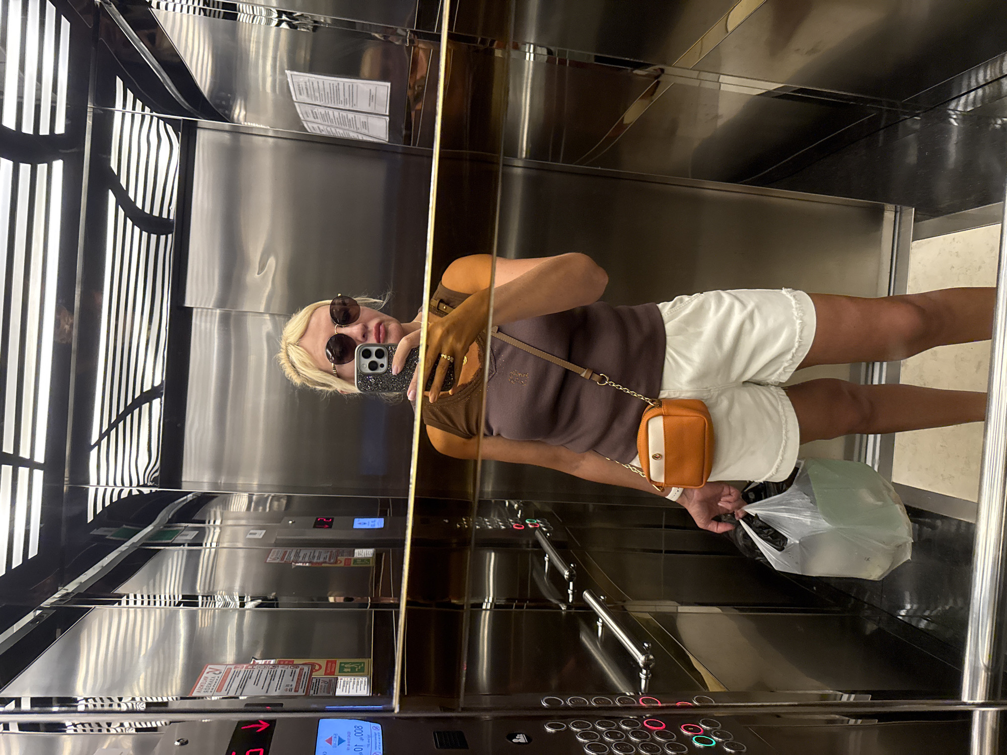 A woman stands in an elevator, taking a selfie. She has blonde hair and is wearing sunglasses, a brown sleeveless top, and white shorts. She is carrying a clear plastic bag in her left hand and a small orange and white crossbody bag. The elevator walls are mirrored stainless steel, reflecting the overhead lights and other parts of the elevator. There are elevator buttons and a digital display visible on the left wall, showing a red arrow pointing down and the number "008". The floor of the elevator appears to be tiled. The scene is consistent with being in an elevator in Alanya, Turkey, a coastal city on the Mediterranean coast of Turkey known for its tourist accommodations.
