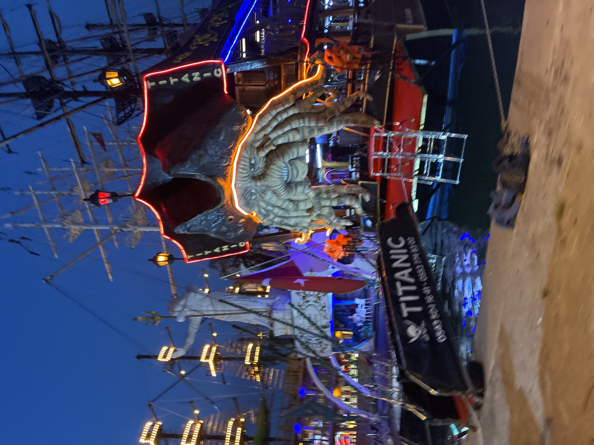 The image shows a nighttime view of a pirate-themed boat docked in Alanya, Turkey, likely in the harbor area. The boat is decorated with numerous lights, including red and blue neon lights outlining its structure and a prominent neon sign spelling out "TITANIC" vertically. A large, grotesque Kraken-like creature sculpture is attached to the side of the boat, with illuminated tentacles. In the background, there are other boats and structures, also illuminated. A statue resembling Neptune holding a trident stands on the boat, with a Turkish flag visible nearby. The boat's name, "TITANIC," is also visible on a banner at the front, along with a phone number. The water reflects the colorful lights. The ground in the foreground is a textured surface, possibly a pier or walkway. No people are clearly identifiable or in focus. The overall scene suggests a tourist attraction or entertainment venue.
