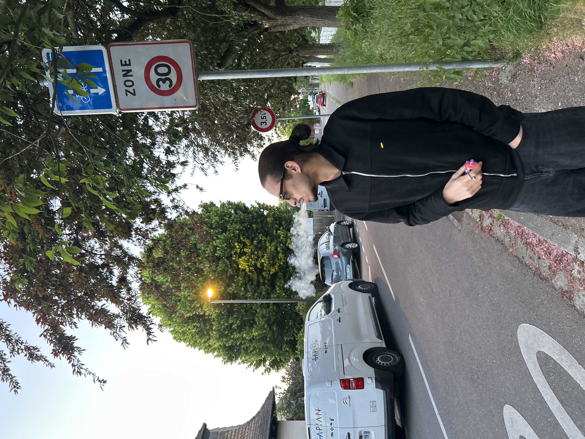 The image shows a man in a black jacket and dark pants, with his dark hair tied in a bun, standing on a street in what appears to be late afternoon or early evening. He is seen in profile, wearing sunglasses and holding a small, colorful vaping device in his left hand, from which a cloud of white vapor is being exhaled from his mouth.
The street is lined with trees and parked cars. Prominently visible on the left are two road signs mounted on a pole: a blue rectangular sign with a white bicycle symbol and an arrow, next to a white square sign with "ZONE 30" written on it, indicating a 30 km/h speed limit zone. Further down the street, another circular red-bordered sign shows "3.5t," likely a weight restriction.
A white Citroën commercial van, with "SAPIAN" and "Agence de Strasbourg" visible on its side, is parked on the right. Behind it, a grey hatchback is visible, with what appears to be exhaust or steam rising from its rear. A street lamp with a warm, yellow glow is illuminated in the mid-ground. Part of a large "30" numeral is painted directly on the asphalt road surface, reinforcing the speed limit. The clear sky and the illuminated street lamp suggest the transition from day to night. The visible text "Agence de Strasbourg" confirms the location context of Strasbourg, France.