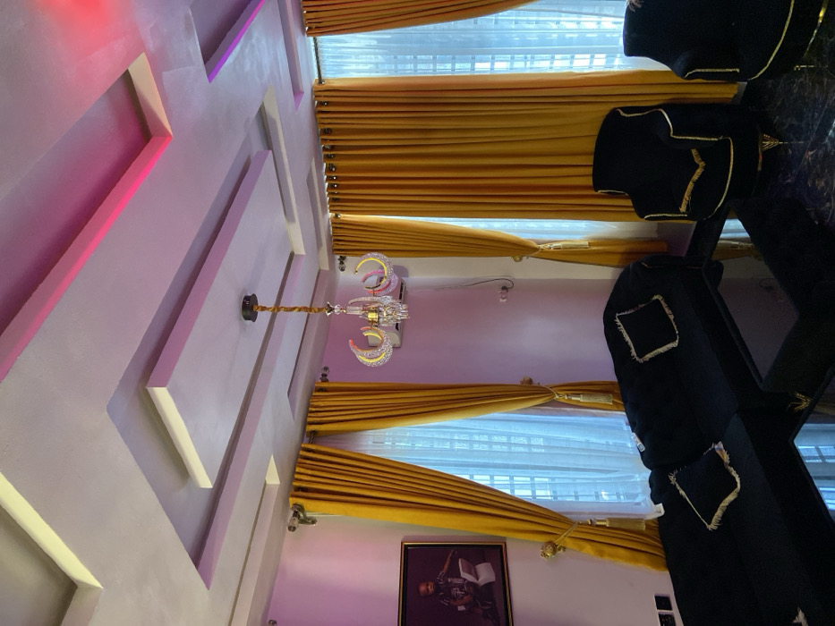 The image displays a modern interior living space situated in Alor, Nigeria. The room features a multi-tiered false ceiling with integrated linear lighting elements emitting pink and red light. A gold-finished chandelier with five crystal-decorated, curved arms hangs centrally. Light-colored walls are flanked by several windows covered with floor-length, golden-yellow curtains secured by gold tassels, revealing sheer white inner drapes. Furniture includes a black sectional sofa with black and gold-trimmed throw pillows, positioned on the left, and at least two black armchairs with gold accent trim and gold-colored legs on the right. A dark, reflective coffee table occupies the central foreground. The floor is covered in dark, reflective tiles. A framed portrait depicting a young boy holding an item resembling a book or tablet hangs on the left wall. An air conditioning unit is mounted on the wall to the right of the chandelier. The scene is devoid of people, actions, or interactions.
