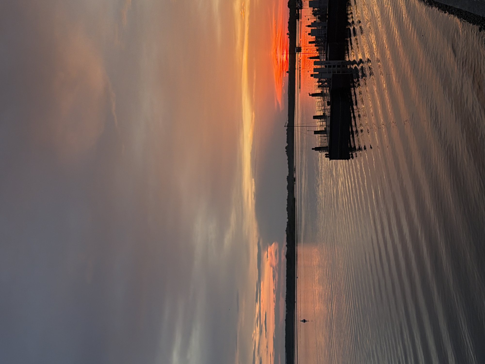 The image depicts a sunset over a body of water, likely a river or lake, with a distant shoreline and a dock in the foreground. The sky is filled with a gradient of colors, ranging from deep grays and purples on the left to vibrant oranges and reds on the right, indicating the sun is setting near the horizon. Clouds are present in the sky, catching the light and adding texture to the scene.
The water is calm, with gentle ripples reflecting the colors of the sky, creating a shimmering effect. A white buoy is visible in the water on the left side of the image. The shoreline in the distance appears forested.
In the foreground, a dock with multiple pylons and railings extends into the water. The dock is silhouetted against the brightly lit sky, with its structure casting reflections on the water. The right edge of the image shows a paved embankment or pathway leading down to the water.
There are no people or discernible actions taking place in the image. The scene is a natural landscape captured during sunset. The location is described as Solikamsk, Russia.