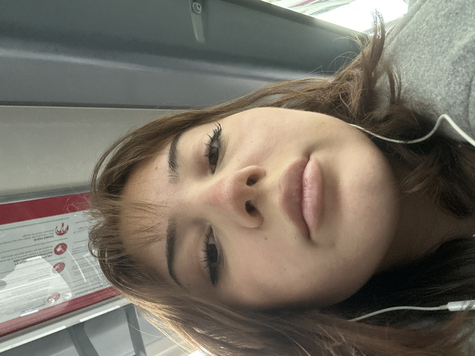 A close-up, low-angle self-portrait features a female individual with light brown hair and bangs, positioned within what appears to be the interior of a public transport vehicle in Schiltigheim, France. Her eyes are directed downwards towards the camera, and her lips are closed. The bridge of her nose exhibits visible redness. She is wearing white wired earphones, with the wires visible extending from beneath her chin. A grey garment, possibly a coat or hoodie, is visible on her left shoulder. In the upper left background, an overhead red and white sign with French text is partially visible. To the upper right, a grey vertical panel with a light-colored handle indicates the vehicle's interior structure.