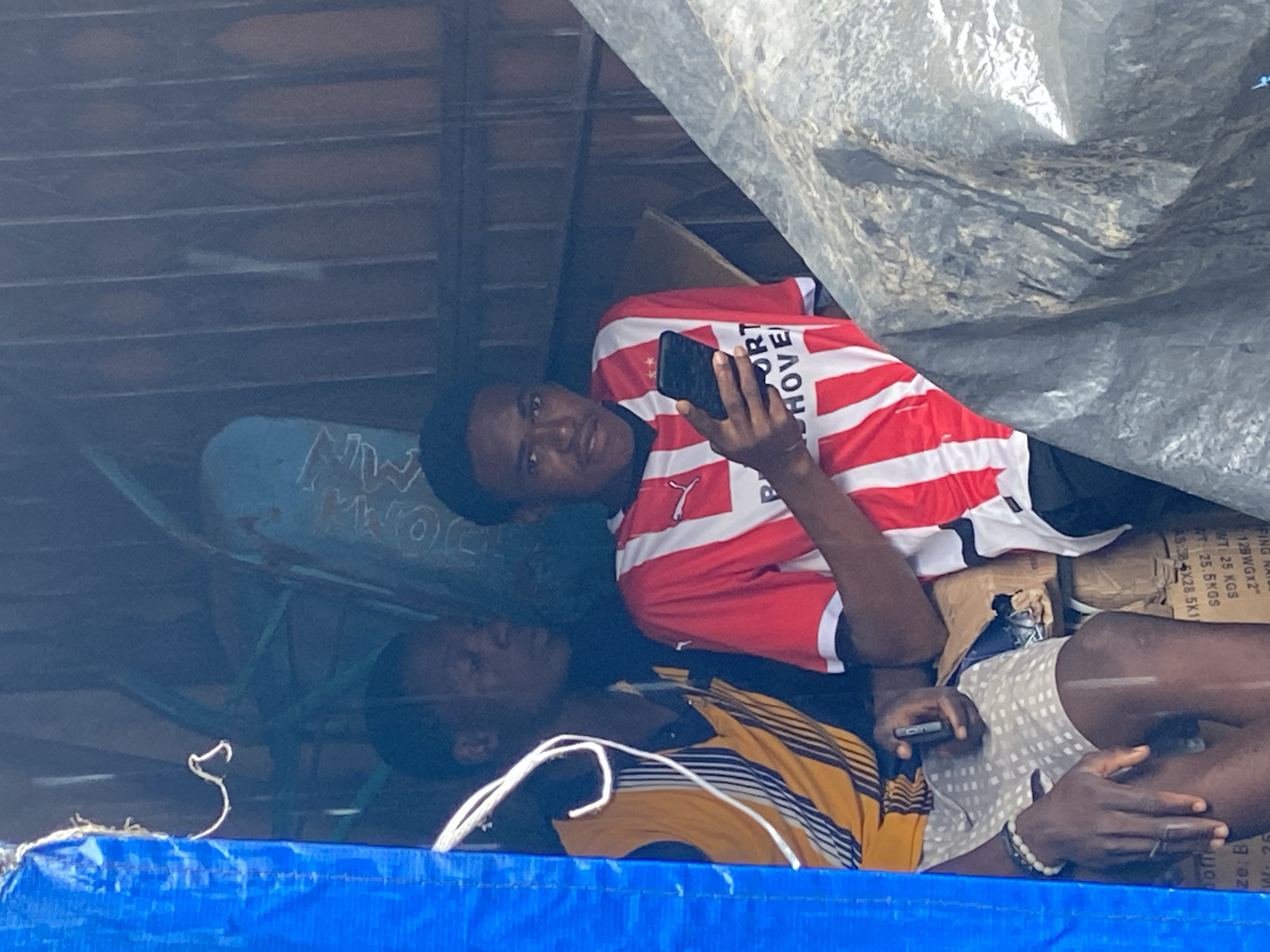 The image shows two young men in what appears to be a crowded, makeshift shelter. The man in the foreground, wearing a red and white striped shirt with "SPORT HOVE" visible on it, is holding a smartphone and looking at it. He has a relaxed posture. Partially visible behind him, another young man wears a yellow and black striped shirt. The shelter is covered with what looks like a large tarp or plastic sheeting, creating a dimly lit environment. Boxes are stacked in the background, some with visible text labels in English, suggesting a possible storage or temporary living situation. A blue tarp is visible in the foreground, suggesting an outdoor or partially exposed setting. The overall impression is one of everyday life in a potentially challenging environment, with the subjects finding moments of leisure or connection through their phones. The text "Ogbunike, Nigeria" in the prompt indicates the geographical context of this scene.