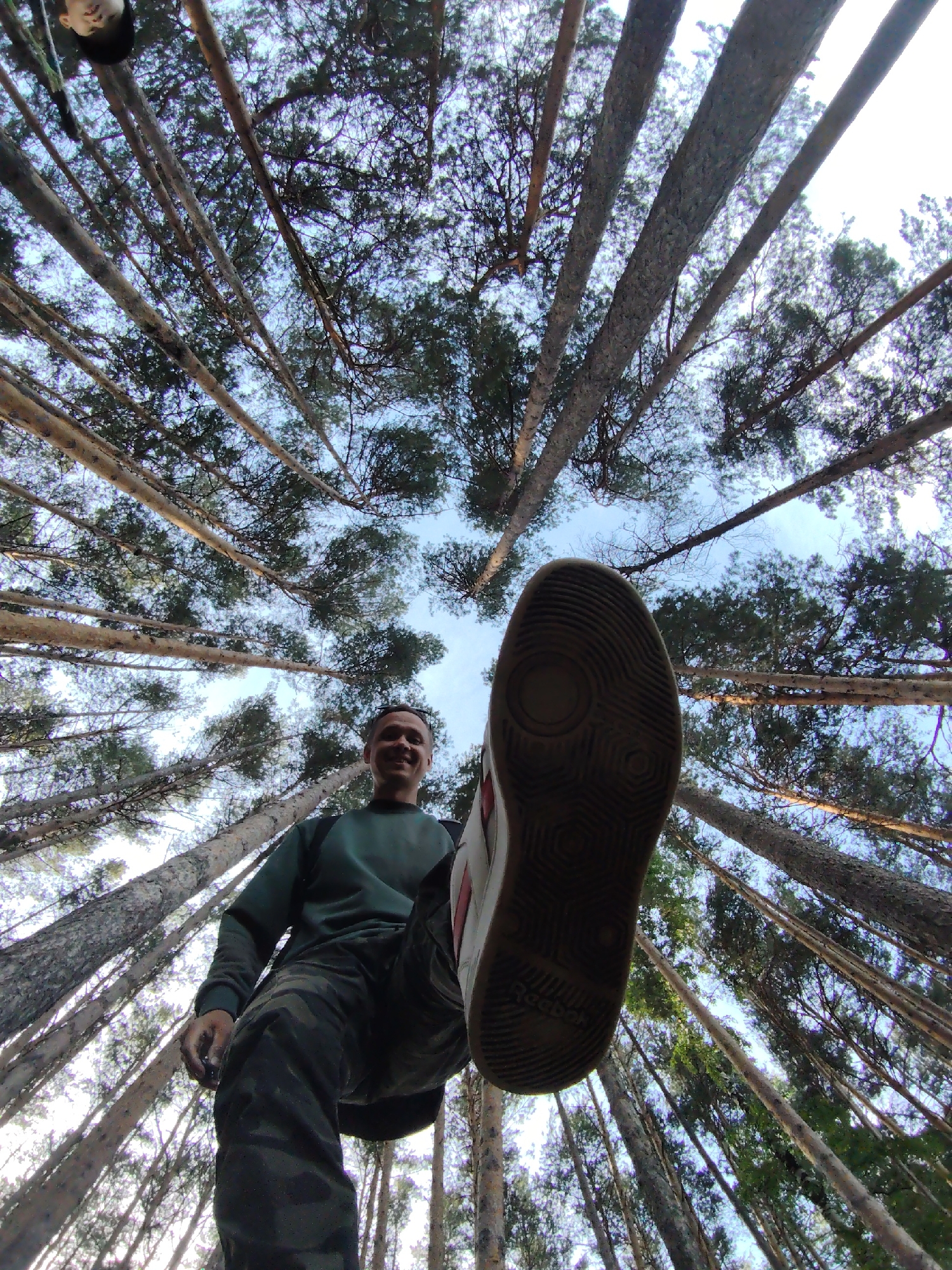 This low-angle photograph captures a dynamic scene within a dense pine forest in Chaikovsky, Russia. The primary subject is an adult male, seen from below as he appears to step directly over the camera. His right shoe sole, bearing the partially visible brand name "Reebok," is prominently featured in the extreme foreground, creating an immersive and somewhat playful perspective.
The man is wearing a dark green long-sleeved shirt and camouflage-patterned pants, with backpack straps visible on his shoulders. He is smiling, suggesting a cheerful or adventurous mood. Surrounding him, numerous tall and slender pine tree trunks ascend steeply, their canopies forming a dense green roof against a clear, bright blue sky. The wide-angle lens used for the shot accentuates the height of the trees and their convergence towards the top of the frame.
The image conveys a sense of outdoor activity, likely hiking or exploring the forest. The bright sky indicates clear weather during daytime, possibly late morning or early afternoon, with light filtering through the tree branches. The overall impression is one of enjoying nature and an active lifestyle within a serene forest setting.
