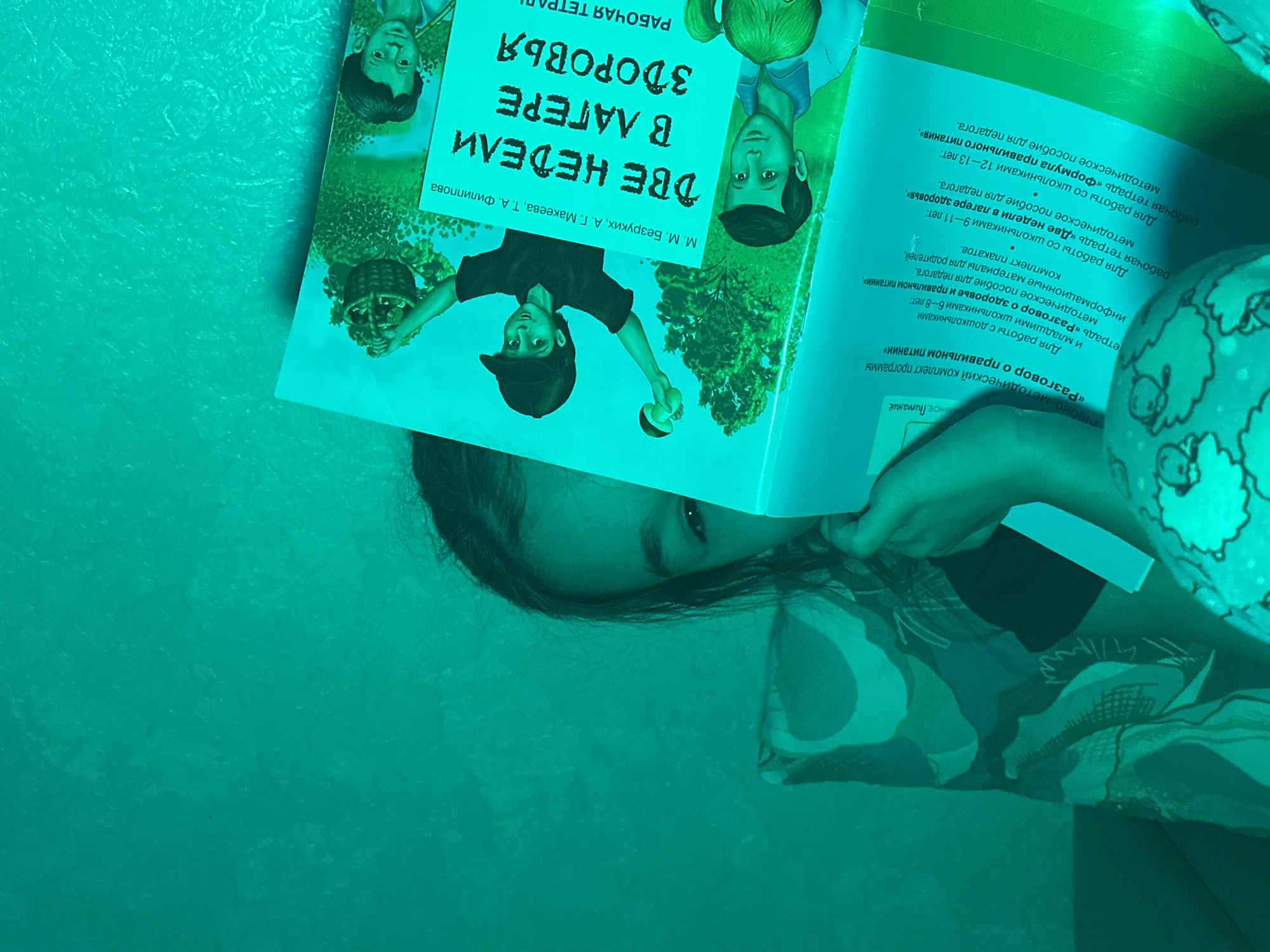 The image captures a close-up indoor scene where a person, likely a child or young girl, is engrossed in reading a book. Only the upper part of her face, including her eyes and forehead with dark hair, is visible, suggesting a focused expression. Her hands are holding the open book, which features a vibrant, light blue cover with illustrations of two boys and green foliage, likely depicting a natural outdoor setting.
The main title on the book cover is "ДВЕ НЕДЕЛИ В ЛАГЕРЕ ЗДОРОВЬЯ" (TWO WEEKS IN A HEALTH CAMP), identified as a "РАБОЧАЯ ТЕТРАДЬ" (WORKBOOK) by authors M. M. Bezruikh, A. G. Makeeva, and T. A. Filippova. Adjacent text describes an "учебно-методический комплект программы «Разговор о правильном питании»" (educational and methodological kit for the program "Conversation about proper nutrition"). This program offers materials for different age groups:
- For 6-8 year olds: "тетрадь «Разговор о здоровье и правильном питании»" (workbook "Conversation about health and proper nutrition"), a methodological guide for teachers, informational materials for parents, and a set of posters.
- For 9-11 year olds: the visible "рабочая тетрадь «Две недели в лагере здоровья»" (workbook "Two weeks in a health camp") and a methodological guide for teachers.
- For 12-13 year olds: "рабочая тетрадь «Формула правильного питания»" (workbook "Formula for proper nutrition") and a methodological guide for teachers.
The background is a plain, greenish-blue surface, possibly a wall or bedding, and a portion of a patterned fabric (likely clothing or a blanket) is visible in the lower right, suggesting a comfortable, relaxed environment. The lighting appears artificial, casting a cool, greenish tint over the scene. The overall activity is a quiet moment of reading and learning. The location context is Uva, Russia.