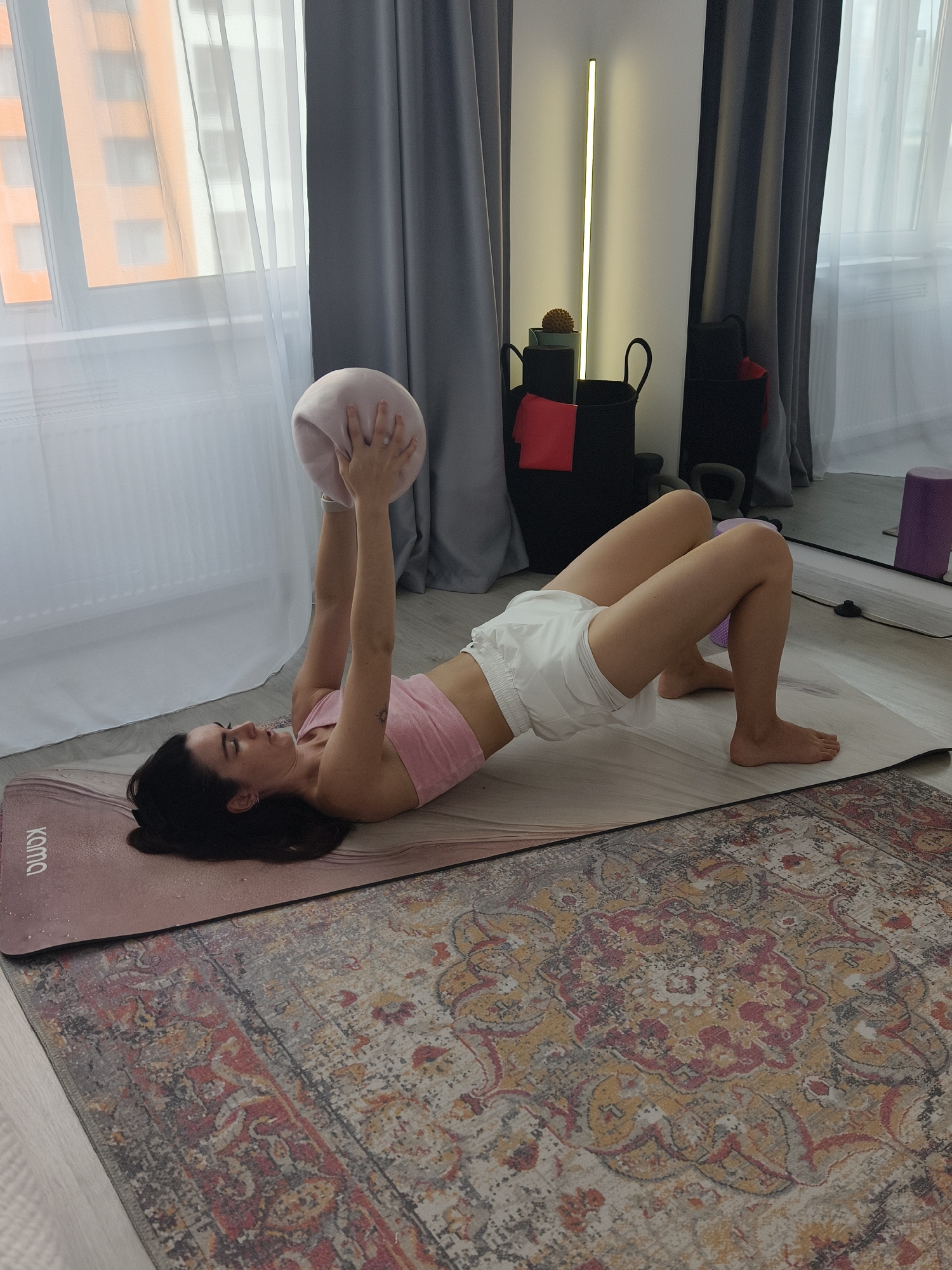 A woman with dark hair lies on her back on a light-colored yoga mat, performing a glute bridge exercise. She wears a pink sports bra and white shorts, holding a light pink exercise ball above her chest with both hands. Her hips are raised, and her feet are planted on the mat, showing a focused yet serene expression with a subtle smile.
The yoga mat, marked with the text "KQMQ" on its visible end, rests on a patterned rug featuring faded red, yellow, and grey designs. The setting is an indoor room, likely a home, with a large window to the left. White sheer curtains and dark grey curtains partially cover the window, through which parts of an orange building are visible, suggesting an urban environment like Tyumen, Russia.
In the background, a tall, slim, vertical LED floor lamp emits a warm glow. Nearby, two black storage baskets contain various items, including a red fabric and a spiky massage ball. Kettlebells and a purple foam roller are also visible on the floor, indicating a dedicated home workout space. The soft daylight entering through the window, combined with the indoor lamp, suggests it could be morning or late afternoon. The scene captures a moment of calm and concentration during a home fitness routine.