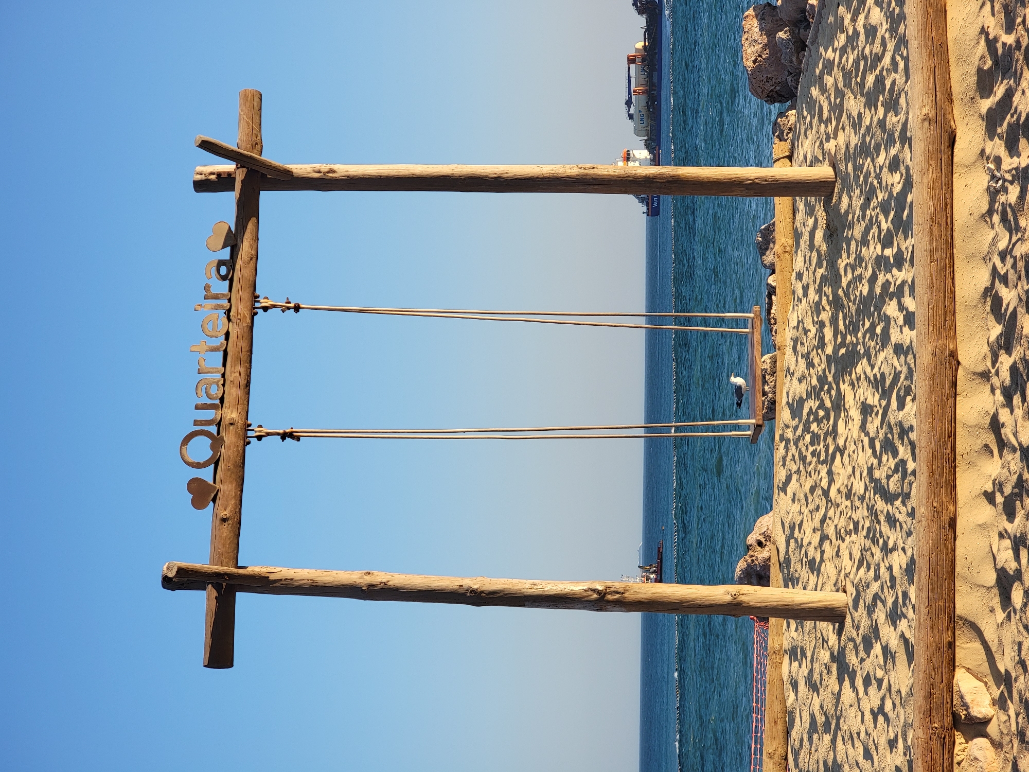 The image shows a wooden swing set on a sandy beach in Quarteira, Portugal. The swing set has the word "Quarteira" with two heart symbols above it. The sea is visible in the background with a ship on the horizon. A seagull is perched on the swing seat. The sky is clear and blue. The sand is textured with footprints and shadows. Rocks and driftwood are scattered on the beach.