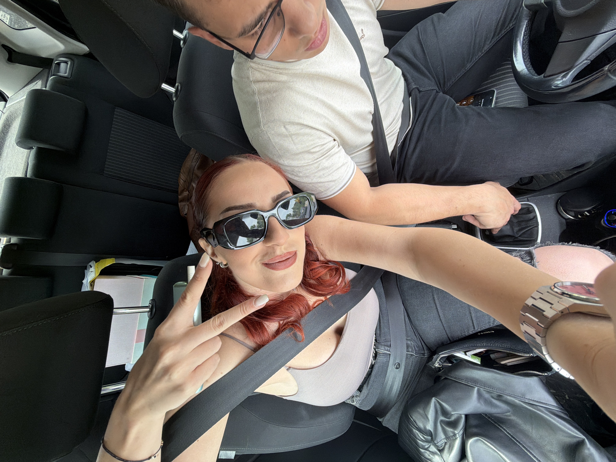 A selfie shows two people inside a car. A young woman with red hair and sunglasses flashes a peace sign. The car is black. The male driver wears glasses and a light-colored shirt. They are both wearing seatbelts. The location is unknown.