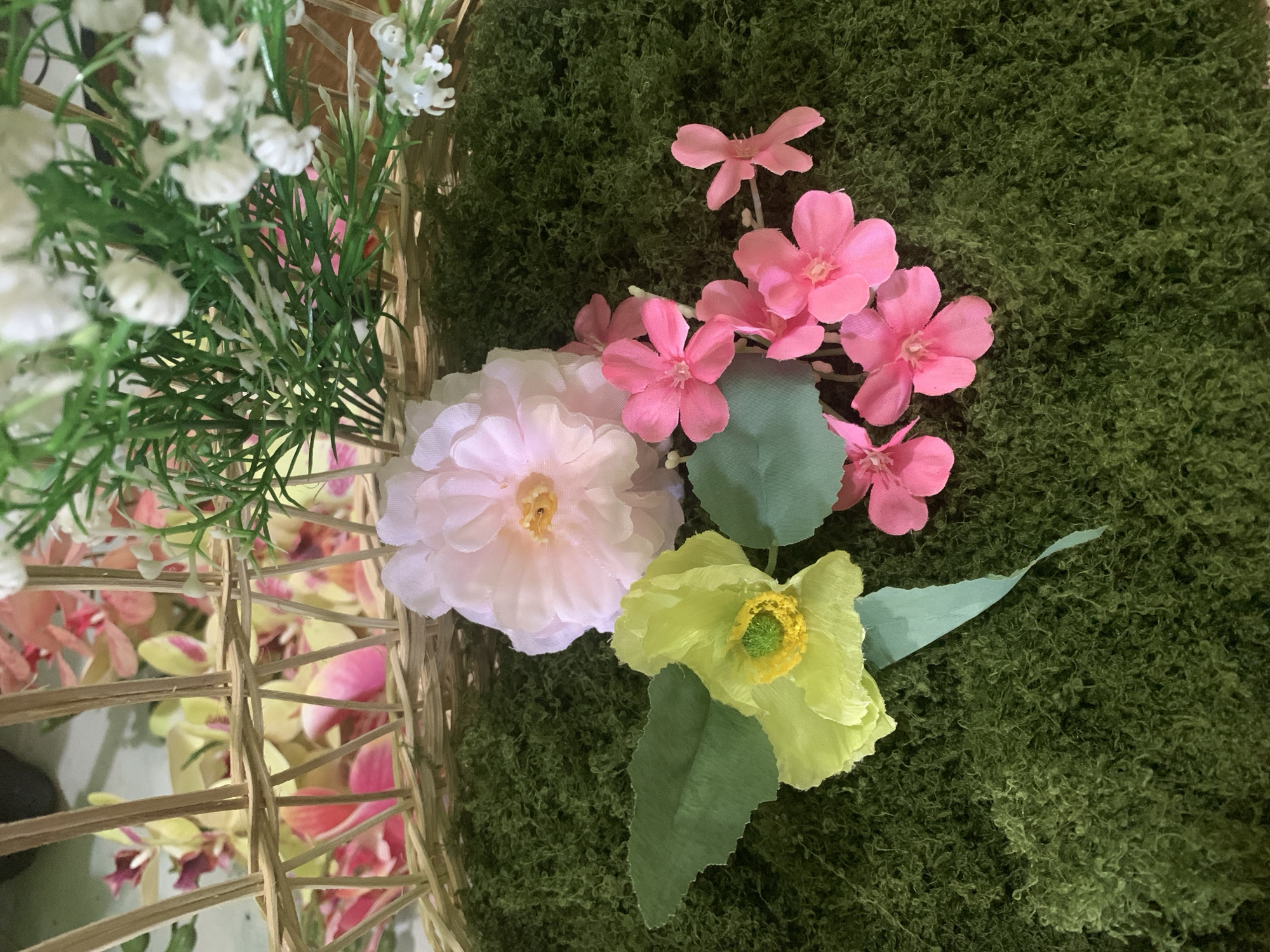 The image displays a close-up, top-down view of artificial floral arrangements set on a bed of green moss. In the foreground, several artificial flowers are arranged: a large, pale pink flower with a yellow center; a bright yellow flower with a green center and two visible green leaves; and a cluster of smaller, vibrant pink flowers. In the background, a woven basket is visible, containing more artificial flowers, including pale pink and yellow orchids, and sprigs of small white flowers with green foliage. The lighting is consistent across the scene, suggesting an indoor setting. There are no people or animals present. The overall impression is one of decorative artistry. The provided information about the location in Kuala Pembuang, Indonesia, cannot be visually confirmed from the image itself.