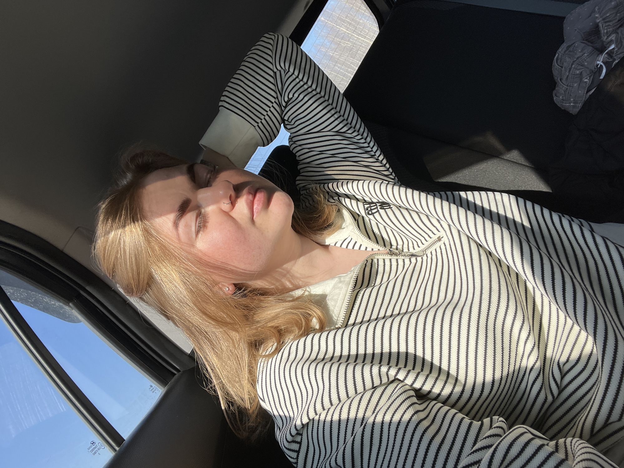 A fair-skinned, blonde-haired woman is reclining in the passenger seat of a vehicle. She is wearing a striped zip-up sweater and has a nose ring. Her head is resting against the window, and her eyes are closed. The interior of the car is visible, including the dashboard and the back seat. Sunlight is streaming into the car through the windows. The location cannot be determined from the provided image.