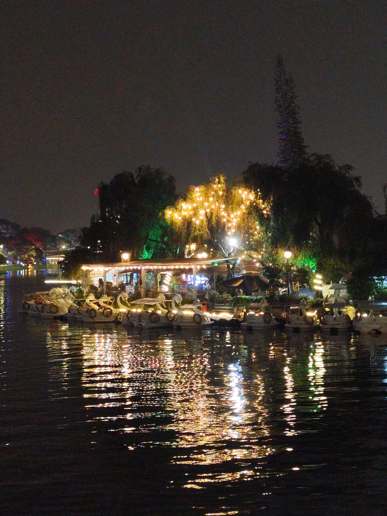The image shows a nighttime view of Xuan Huong Lake in Da Lat, Vietnam. Several boats, appearing to be swan-shaped paddle boats, are docked. The shoreline is illuminated with various lights and decorations. Trees are visible, with one particularly large tree adorned with many small, warm-toned lights. The lights reflect in the water, creating streaks and patterns on the lake's surface. The sky is dark and indistinct.