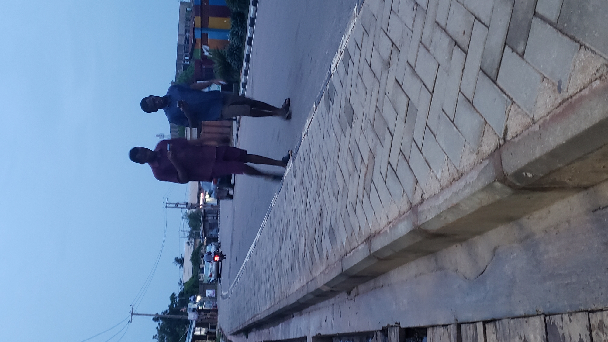 An outdoor image, captured at what appears to be dusk or early evening, shows a street scene in Narayi, Nigeria. The sky is a muted blue-grey. The photograph is oriented vertically, showing the street and sidewalk at an angle.
In the foreground and mid-ground, two men are visible, seemingly walking or standing near the edge of a paved road. The man on the left wears a dark blue short-sleeved shirt and dark shorts. The man on the right wears a dark red/maroon short-sleeved shirt and dark shorts, holding a small white object, possibly a phone. Both men have dark complexions. Their legs and feet show some motion blur, suggesting movement.
To the right, a raised sidewalk or pedestrian path is prominent, constructed from light-colored paver blocks arranged in a herringbone pattern. A concrete curb separates this path from the asphalt road. In the background, other vehicles including cars and a motorbike are partially visible, along with various buildings and structures. Some buildings in the far distance feature colorful panels, potentially shipping containers or distinct architectural elements. Utility poles and overhead wires crisscross the sky on the left side of the frame. No discernible text is visible anywhere in the image.