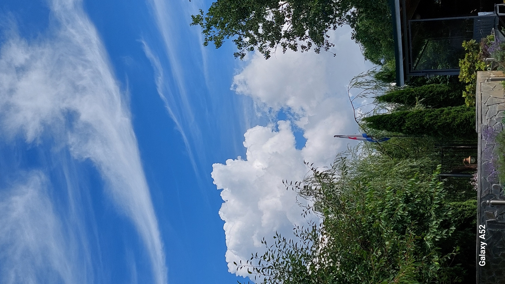 This image captures a view of the sky and foliage in Zhavoronki, Russia. The sky is a vibrant blue, dotted with wispy white cirrus clouds and larger, fluffy cumulus clouds. In the foreground, lush green trees and shrubs are visible, with some foliage drooping. A flag, appearing to be the Dutch flag with its red, white, and blue horizontal stripes, flies from a flagpole. Several conical evergreen trees stand behind the other vegetation. A dark, modern-looking structure with a mirrored or reflective surface is partially visible on the right side of the frame. The bottom of the image shows a stone wall. The "Galaxy A52" watermark is present in the lower left corner.
