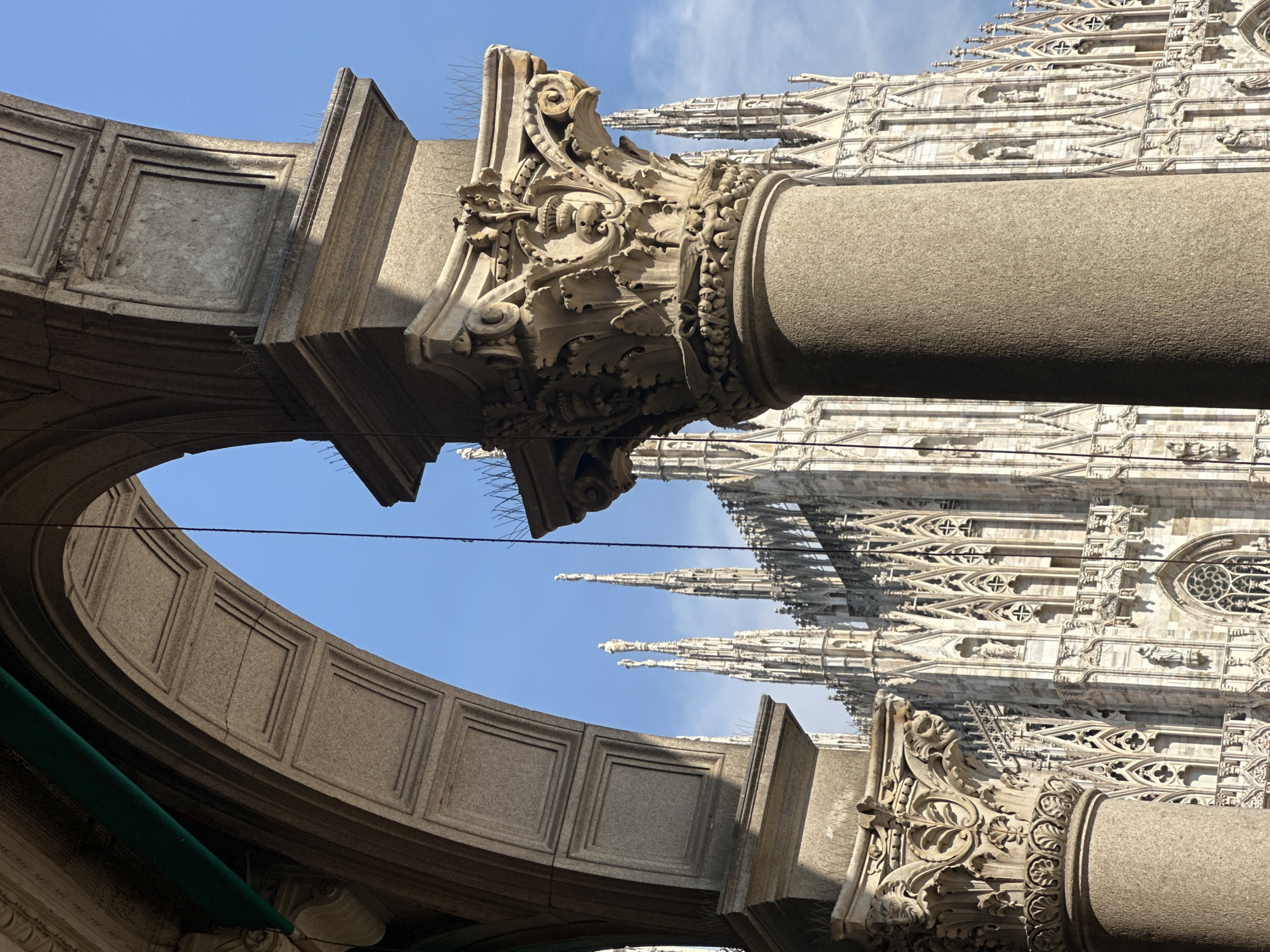 This is a photograph of the Milan Cathedral (Duomo di Milano) in Milan, Italy, as seen through an architectural archway and framed by columns. The cathedral's ornate Gothic spires and facades are visible in the background, under a clear blue sky. In the foreground, a large stone column with intricate carvings is positioned to the right, partially obscuring the view of the cathedral. Another column and part of an arched structure are visible on the left. The image captures a daytime scene, with sunlight casting shadows on the architectural elements. There are no people visible in the photograph.