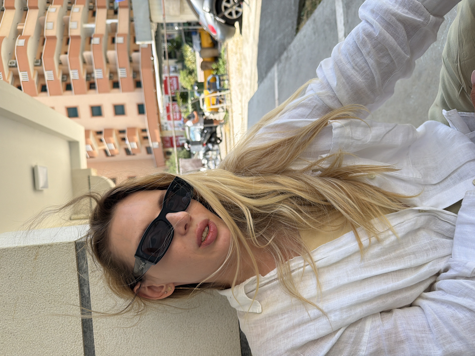 The image shows a young woman with long blonde hair wearing a white linen shirt and sunglasses. She is sitting in front of a building with balconies and an open-air area. In the background, there are more buildings, a street with parked cars and scooters, and a few people. The scene appears to be in a city, possibly Budva, Montenegro, given the architectural style of the buildings. The woman's hair is being blown by the wind.