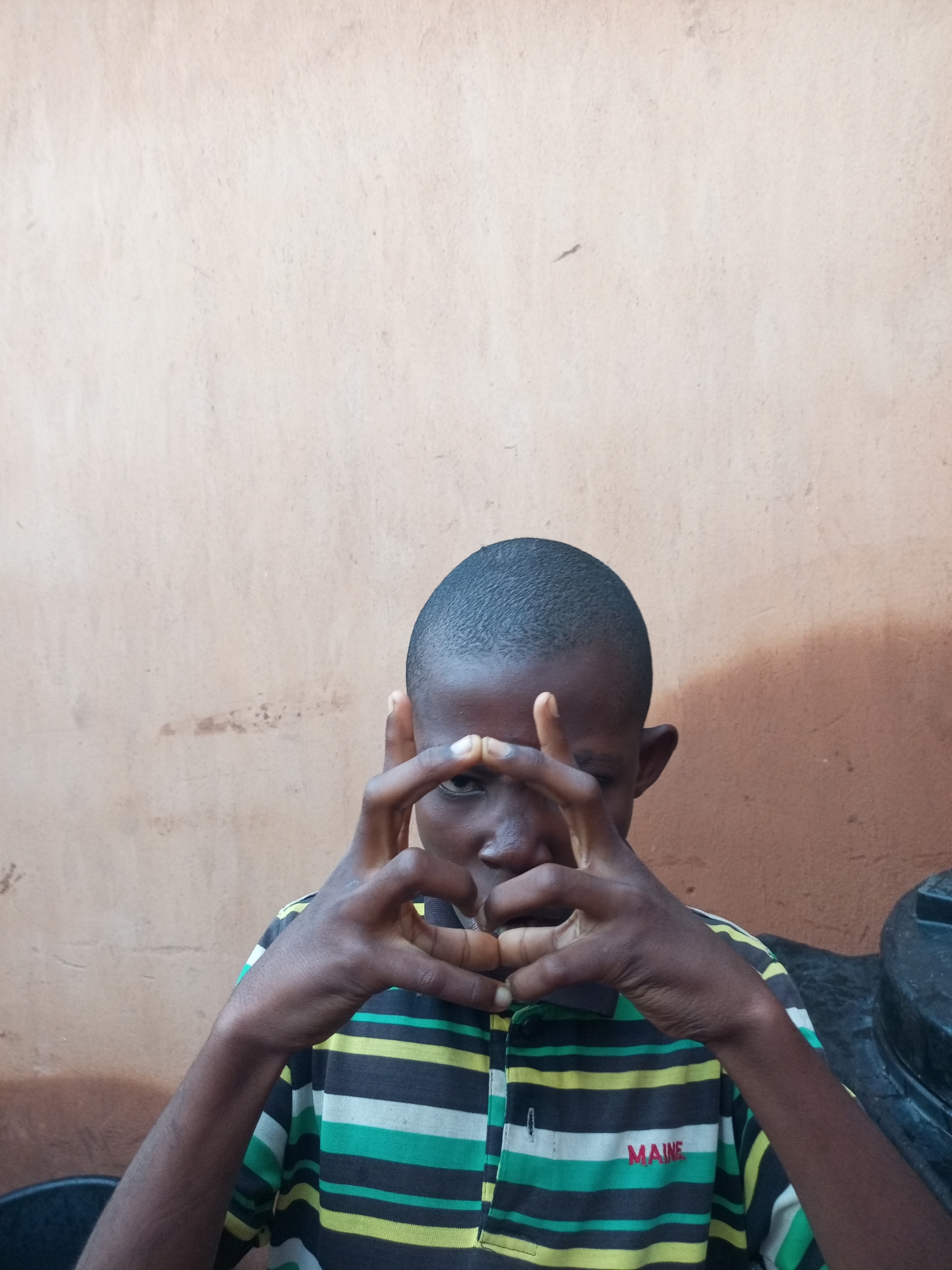 A young boy with dark skin and short hair is pictured from the chest up, in front of a textured, light brown wall. He is wearing a horizontally striped t-shirt with black, green, yellow, and white bands. His hands are raised in front of his face, with his fingers interlaced to form two circles. The boy is looking directly at the camera. A large black object, possibly a water tank, is visible to his right. The background wall has some dark smudges and a lighter patch towards the top.