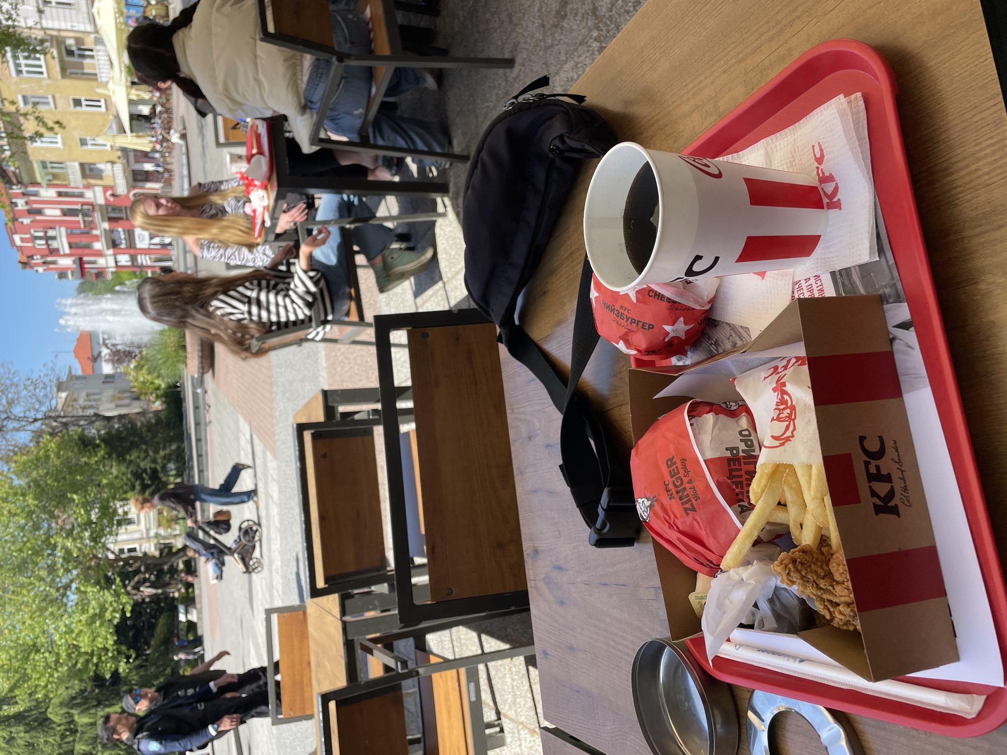 This image captures an outdoor dining scene at a KFC restaurant, likely located on a public square or pedestrian street in Plovdiv, Bulgaria, given the architectural style and Cyrillic text. The time of day appears to be bright and sunny, indicated by clear skies and well-lit areas, suggesting a pleasant afternoon.
In the immediate foreground, a wooden table holds a red KFC tray laden with a fast-food meal. Objects on the tray include a white KFC cup filled with a dark beverage, a KFC Cheezburger wrapped in red paper with Cyrillic text "ЧИЙЗБУРГЕР," and a KFC Zinger burger, also wrapped and labeled "Hot & Spicy." A carton of french fries and a piece of fried chicken are visible in an open KFC box. KFC branded napkins are also present. Next to the tray, a black fanny pack or sling bag rests on the table. Two round, metallic containers, possibly for sauces or waste, are visible towards the bottom left.
In the mid-ground and background, several people are seated at other outdoor tables, mostly seen from behind or the side. Two women with long hair, one blonde and one dark-haired, are clearly visible, seated opposite each other. Beyond them, a bustling public space unfolds, with other individuals walking. A prominent white fountain can be seen in the distance, indicating a public square. Buildings with traditional European architecture line the street, and green trees add to the urban park-like setting. The general activity is people enjoying meals outdoors and strolling through the city. Visible text includes "KFC" branding, "ЧИЙЗБУРГЕР," "ZINGER Hot & Spicy," "Col. Harland Sanders," and partial Cyrillic words such as "РЕЦЕНА," "ОРИГИ," "АЧЕС," "А ПРЯ," and "ИСТИН."