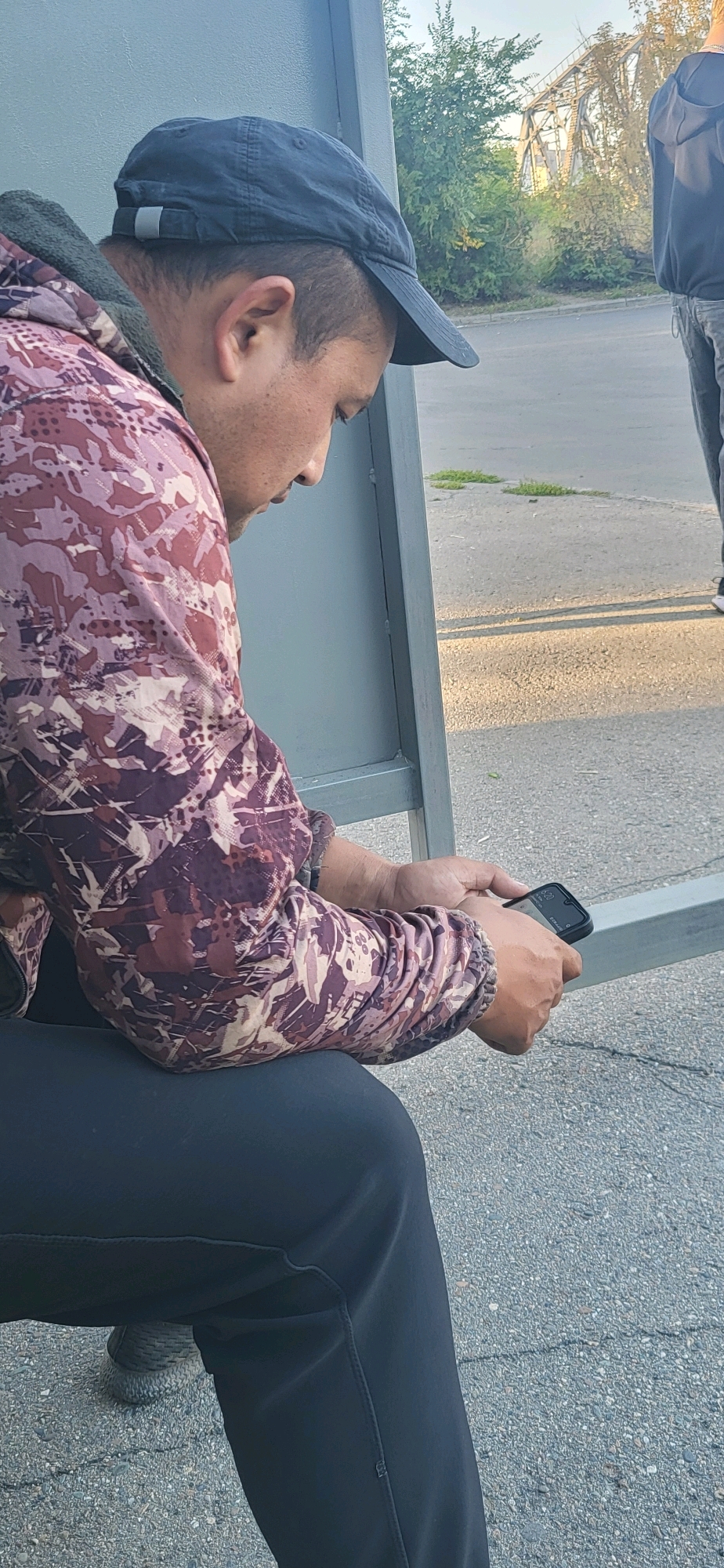A person wearing a dark baseball cap, a multi-colored camouflage jacket, and dark pants is sitting on a bench and looking at a smartphone. The person's head is bowed, and their gaze is directed downwards at the phone. The phone is black and held in the person's right hand. The background shows a gray structure, likely part of the bench or a shelter, and an asphalt surface with scattered patches of grass. In the distance, there are trees and a metal bridge structure. The scene appears to be outdoors in an urban or semi-urban environment.