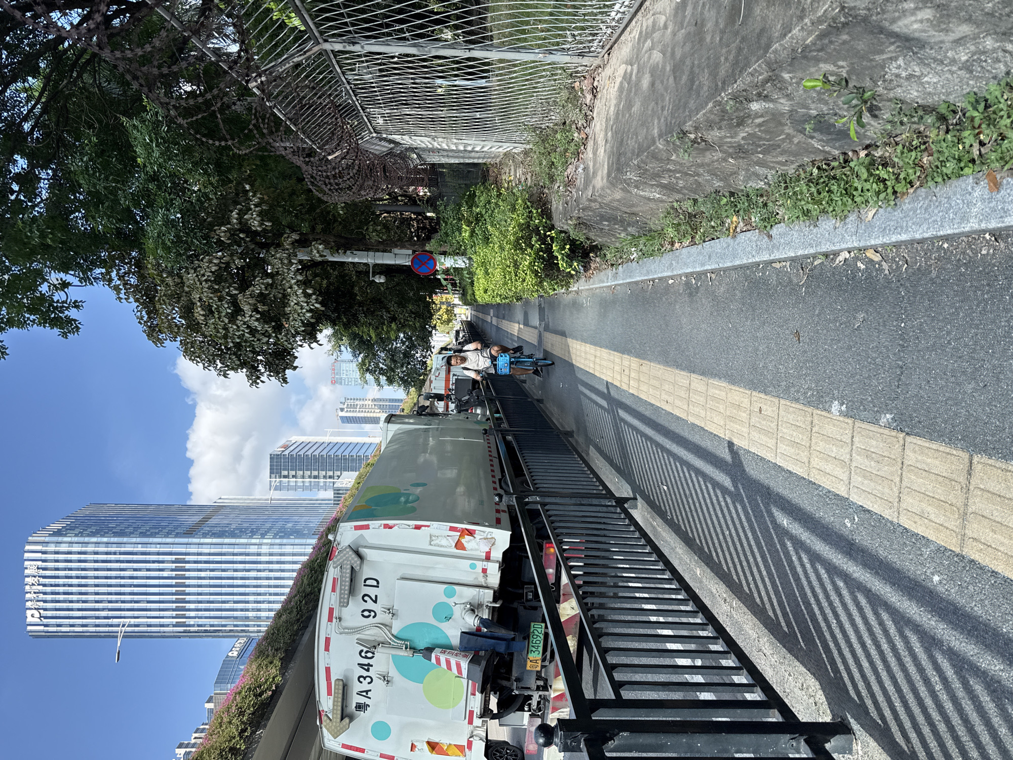 A cyclist rides a blue bicycle on a paved pathway in the Huangpu District, China. The pathway is bordered by a black metal fence on the left and a concrete embankment with vegetation on the right. In the background, a white garbage truck with colorful circular designs is parked on the adjacent road. Beyond the truck, modern high-rise buildings with reflective glass facades dominate the cityscape. The sky is clear with scattered white clouds. A street light and a traffic sign are visible along the pathway. The scene is illuminated by sunlight, casting shadows from the fence onto the pathway.