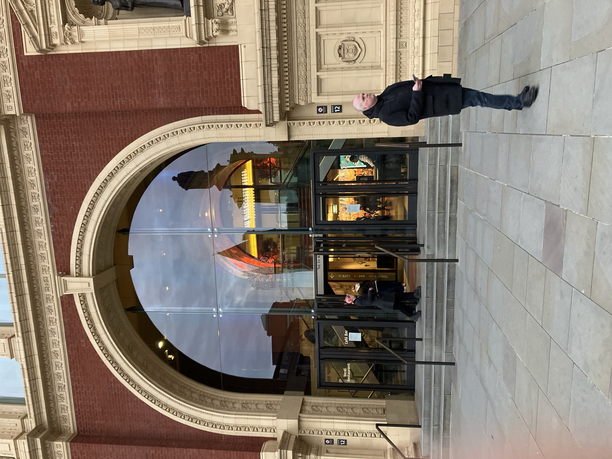 This image captures the exterior of the South Entrance (Door 12) of the Royal Albert Hall, located in South Kensington, London, United Kingdom. The building features a distinctive red brick facade with ornate cream-colored stone detailing, including decorative friezes and a recessed niche containing a dark statue on the right.
The central feature is a large, arched glass entrance comprising multiple panels, with several glass-paned doors at ground level. Text on the doors identifies "Verdi Restaurant" and "Café Bar." A small black sign on the stone wall to the left of the entrance reads "DOOR 12." Another notice, visible through the glass on the right, states: "The South Foyer was opened by Her Majesty The Queen on 30th March 2004."
A series of wide, shallow stone steps, flanked by dark metal handrails, leads up to the entrance from a paved pedestrian area. The paving consists of large, light grey rectangular flagstones.
In the foreground, to the right of the entrance, a bald Caucasian male, dressed in a black hooded jacket and dark blue jeans, stands facing left, holding a smartphone in his right hand.
To the left of the entrance, a person with pink hair, wearing a long dark coat, is seen walking towards or through one of the glass doors. Another individual is partially visible immediately behind them, near the entrance. Through the reflective glass, interior lighting, a staircase, and additional figures are discernible. The glass also reflects external elements, including the silhouette of a statue and surrounding buildings.