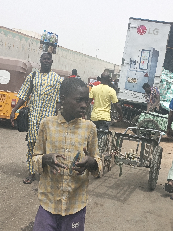 The image captures an active street scene in Fagge C, Nigeria, under bright, diffused daylight.
In the foreground, slightly to the left, a young male with dark skin and short dark hair wears a yellow and grey plaid long-sleeved button-up shirt and purple athletic trousers with "SPORT" visible on the left thigh. He holds a small, round, iridescent object in his left hand, while his right hand is open, gesturing slightly. His gaze is directed towards the right foreground.
Directly behind him, an adult male with dark skin walks towards the right, attired in a blue and yellow plaid short-sleeved tunic and matching trousers. He carries a stack of approximately eight plastic water bottles, secured with black straps, balanced on his head, and a black messenger bag over his left shoulder. Yellow three-wheeled auto-rickshaws are partially visible on the far left.
In the mid-ground to the right, a large white delivery truck is prominent, featuring an "LG" logo and an advertisement for an LG refrigerator on its side. Text on the truck reads "FOUANI NIGERIA L. LTD. NO. 8, NADIN ROAD, KANO STATE" and "TEL: 08061611100." An adult male in a multi-colored plaid shirt is bent over at the rear of the truck, engaged in handling light-colored sacks or bundles. Adjacent to the truck, two two-wheeled handcarts are present on the paved surface; one appears empty, and the other holds some materials. Further back, another adult male, wearing a plain yellow t-shirt, walks away from the camera.
The background features a light-colored concrete wall with a green and brown geometric or floral pattern extending horizontally. Indistinct figures and structures are visible beyond the wall. The ground consists of a dark, worn paved surface. The environment indicates a bustling urban commercial area.