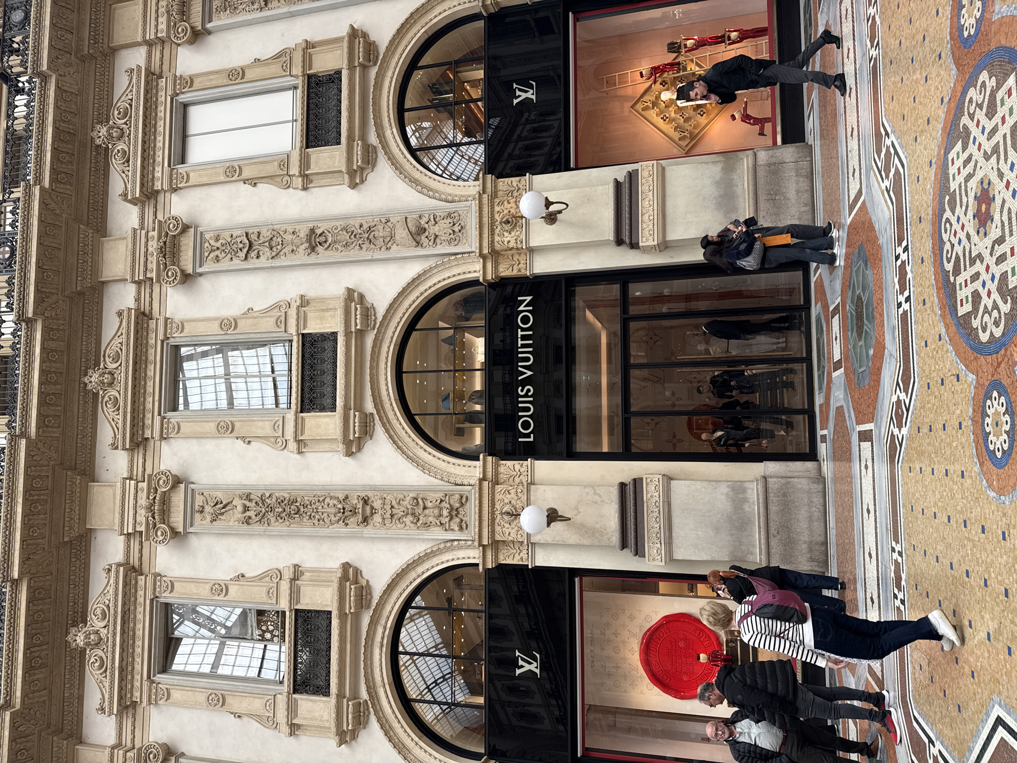 The image shows the Galleria Vittorio Emanuele II in Milan, Italy. The architecture features ornate carvings and arches, with Louis Vuitton storefronts occupying several bays. People are seen walking and looking at the displays. The floor is a mosaic tile pattern.