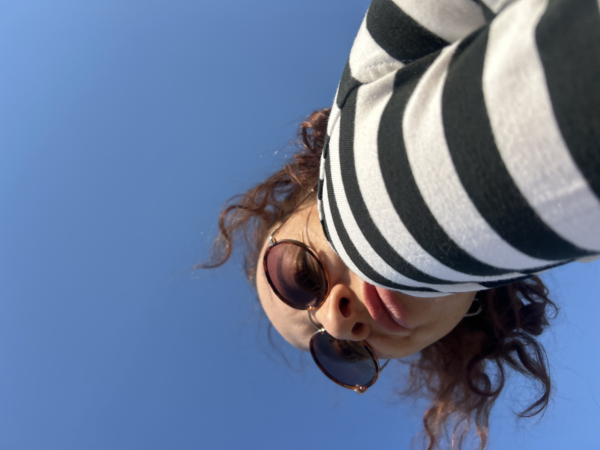 A low-angle, close-up shot looking up at a person with curly brown hair. They are wearing sunglasses with a brown frame and dark lenses. The person is wearing a black and white striped top. The background is a clear, bright blue sky. There are no obvious clues to place the image in Chaikovsky, Russia; the sky and clothing are not specific to that location.