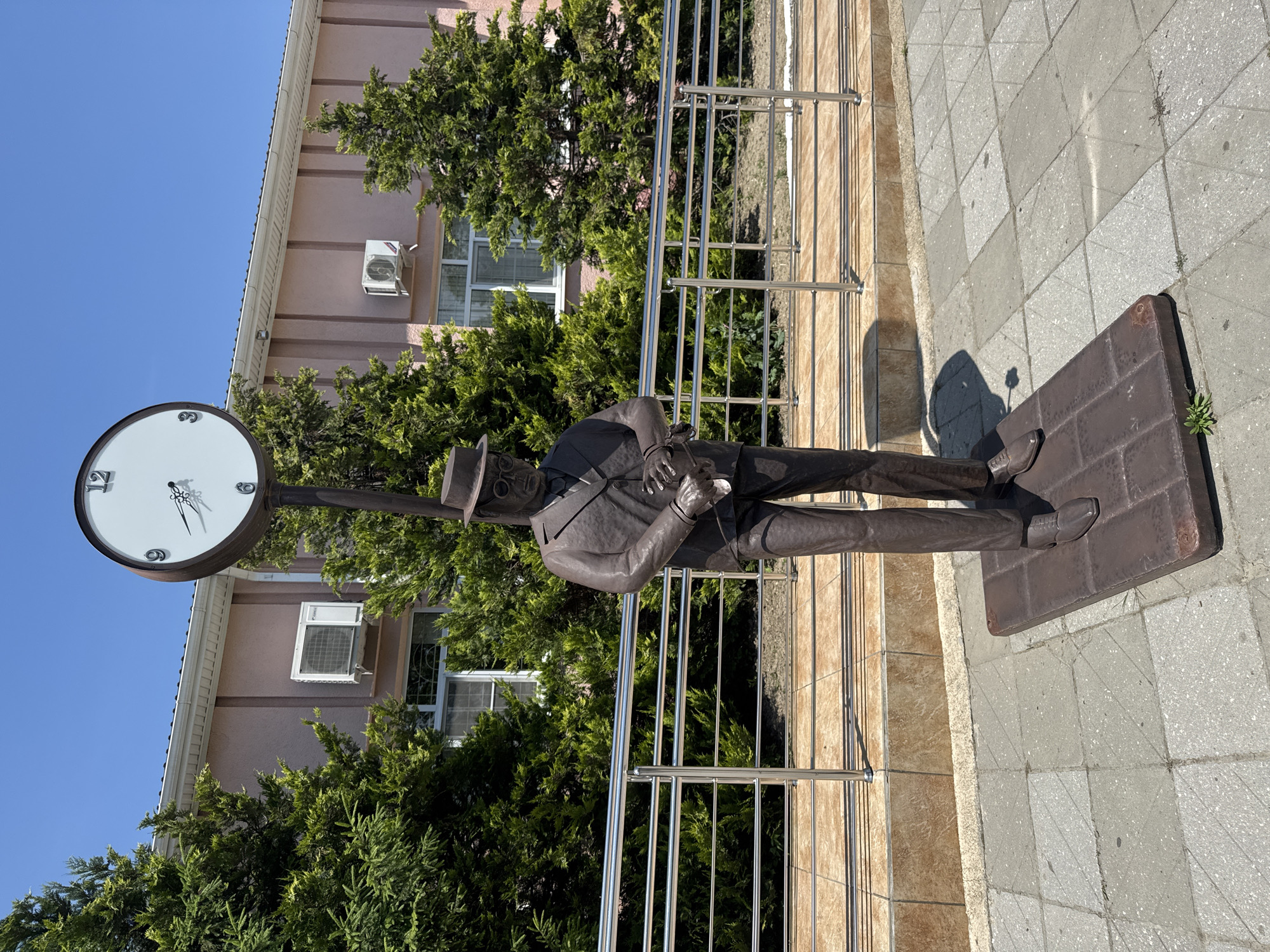 In Slavyansk-na-Kubani, Russia, a bronze statue of a man in a suit and hat stands on a dark rectangular base. The statue is positioned outdoors on a paved walkway, with a building in the background. The building has a light pink facade, white trim, and several windows. Green trees and bushes are visible around the statue and in front of the building. Above the statue, a large clock with Roman numerals and thin black hands is mounted on a pole. The clock face is white with a dark circular rim. The sky is clear and blue. A metal railing runs horizontally behind the statue, separating it from the building. The ground is paved with light-colored square tiles with some dark grout lines. The statue appears to be looking down at its hands, as if checking a watch.