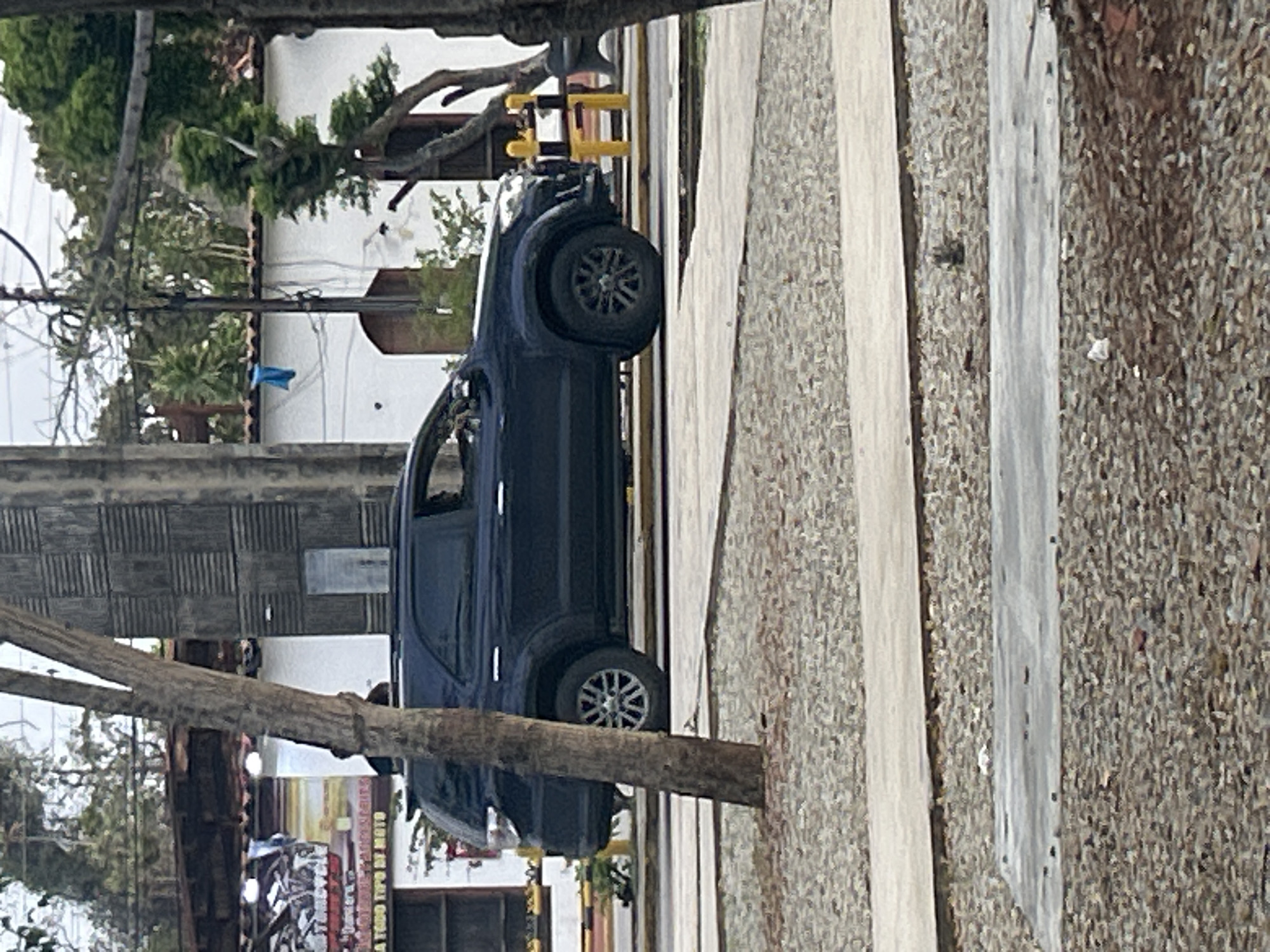The image shows a dark blue SUV parked on a street in Santa Ana, Venezuela. The vehicle is positioned in the foreground, with its side facing the viewer. The surrounding environment includes trees, buildings, and a rocky, gravel-covered area. A wooden pole with a blue cloth draped over it is visible in the background. The sky appears overcast.