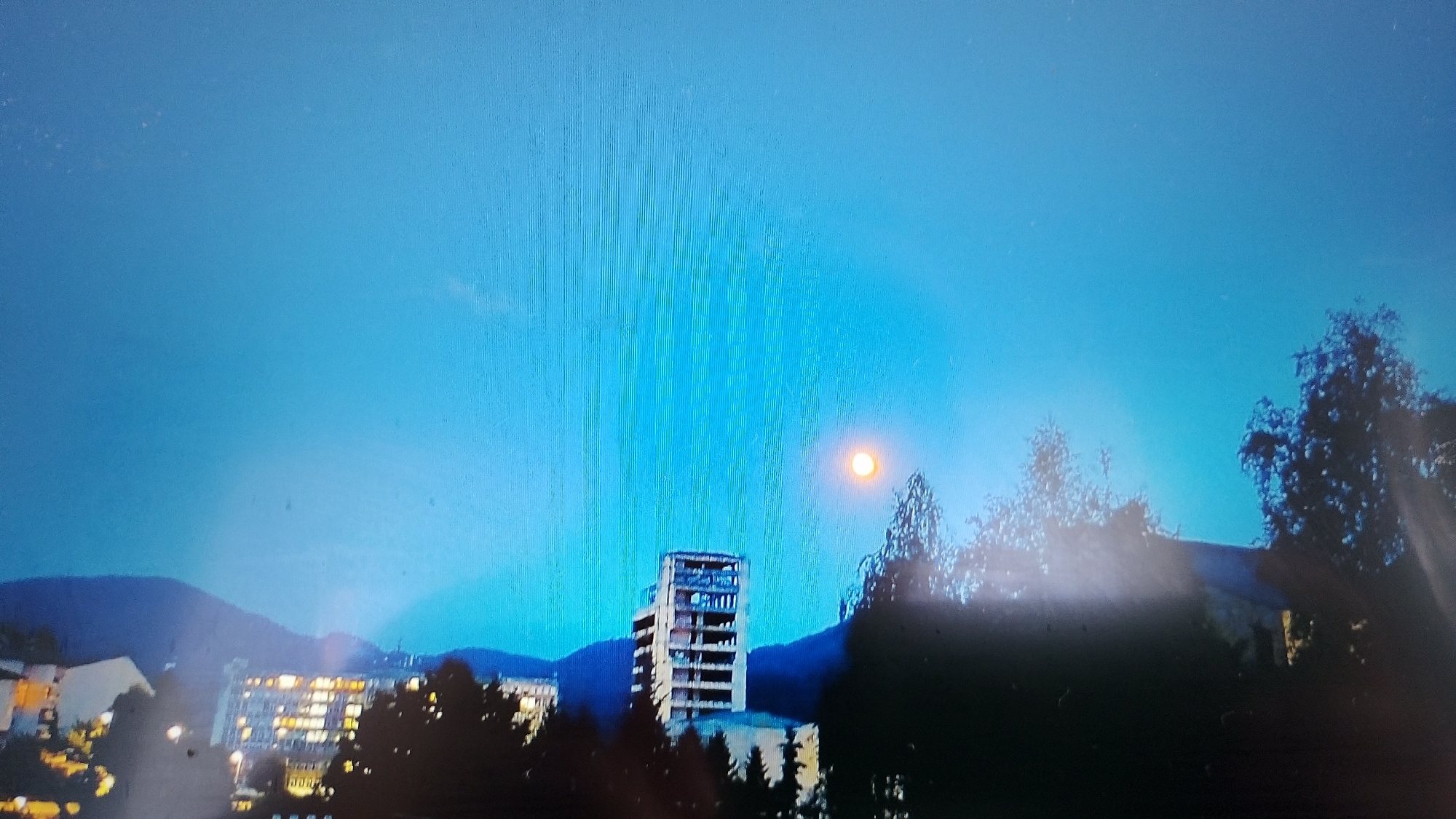 The image depicts a nighttime or twilight cityscape with a large, unfinished concrete building prominently featured. The building appears to be a skeletal structure, with open floors and visible rebar. It is surrounded by trees and other buildings. The sky is a deep blue, with a bright, illuminated object that resembles the moon or a full moon visible in the upper right quadrant of the image. The overall impression is one of urban development and possibly abandonment or a construction site. The image quality is poor, with visible lines and artifacts that suggest it may have been taken with a low-resolution camera or is a photograph of a screen.