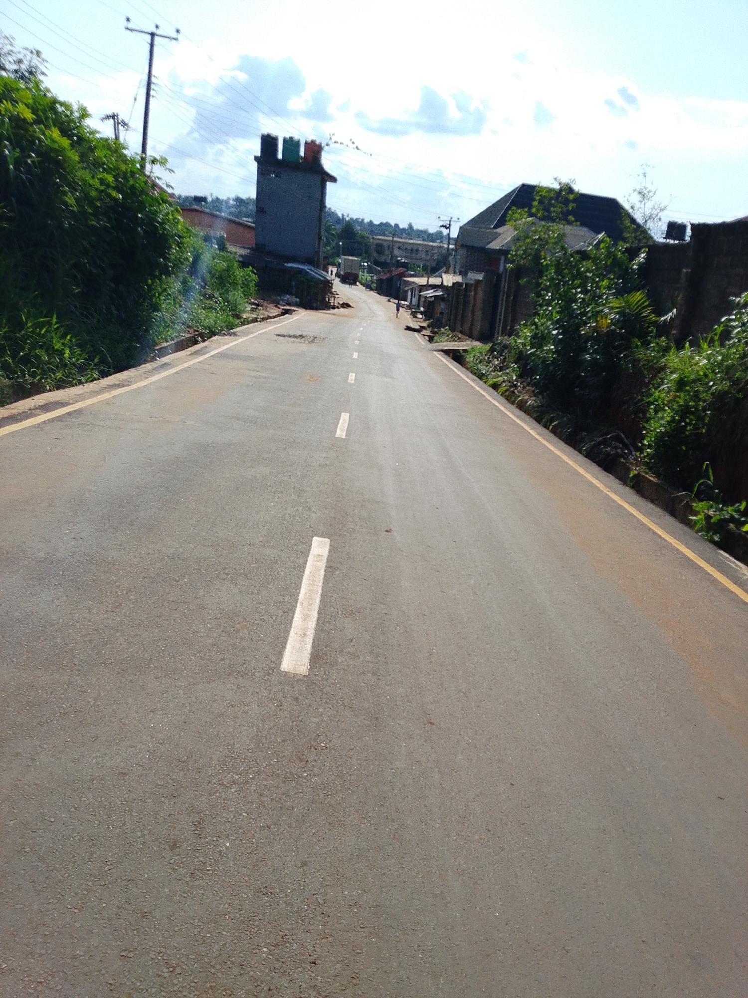 The image shows a straight, paved road in Umuahia, Nigeria, heading uphill into the distance. The road has a yellow line marking the edges and broken white lines down the center. Vegetation lines both sides of the road, becoming denser towards the left. On the left is a gray concrete structure with multiple water tanks on top. Further down the road, a few buildings line both sides. A single person is walking along the right side of the road in the distance. Utility poles and wires are visible in the background under a partly cloudy sky.