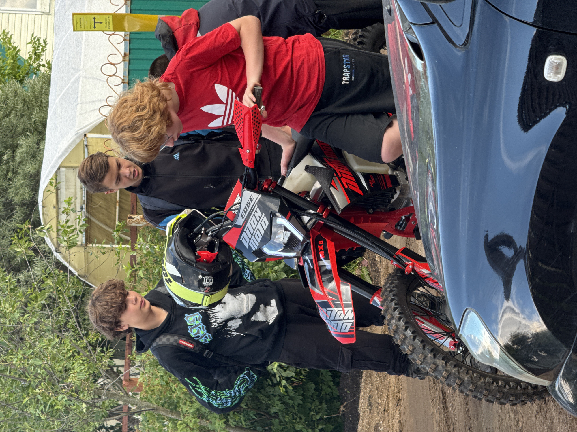 In an outdoor setting, possibly a backyard or a rural area, three young males are gathered around a dirt bike. The male on the left, wearing a black hoodie and black cargo pants, has curly brown hair and is looking down. He has a backpack on. To his right, a male with neatly styled brown hair wears a black athletic jacket with blue accents. He is looking towards the dirt bike. The male in the center-right has curly blonde hair and is wearing a red t-shirt with white graphics and black shorts. He is leaning on the dirt bike and looking down at it. The dirt bike is red and black with white and red decals. A helmet with yellow and black accents is placed on the handlebars. In the foreground, the hood and headlight of a dark-colored car are visible, partially obscuring the front wheel of the dirt bike. The background shows lush green foliage, a fence with a white canopy structure behind it, and a yellow sign with black text and a symbol.
