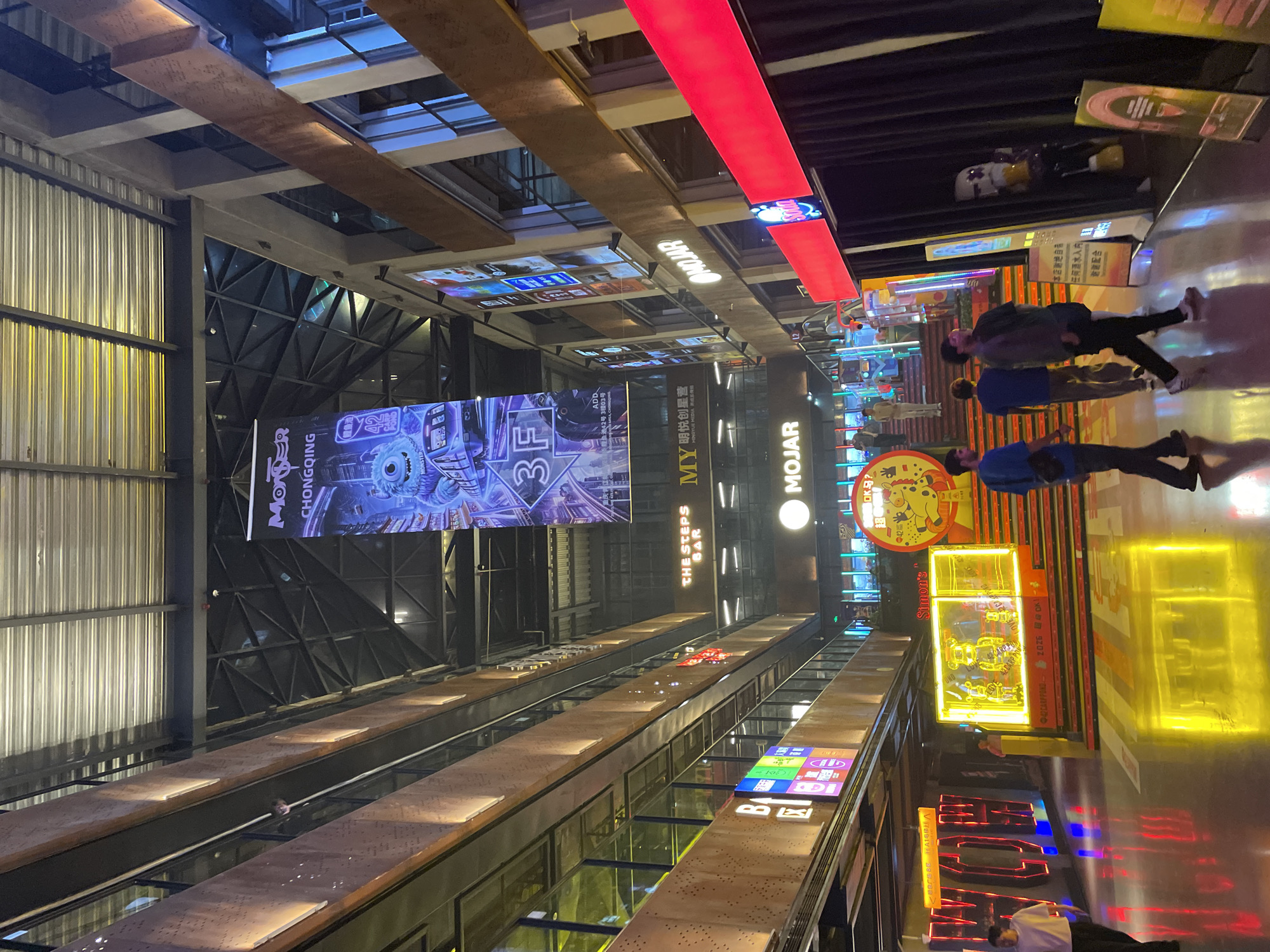 The image shows a bustling indoor entertainment complex in Liangjiang, China. A large vertical banner for "MONSTER CHONGQING 3F" hangs from the ceiling, featuring animated characters and text. Multiple levels of walkways, lined with shops and illuminated signs, create a vibrant atmosphere. In the foreground, people walk on a reflective floor that mirrors the colorful lights. One section features a bright yellow illuminated display with cartoon figures. To the right, a dark curtained entrance and signs suggest a venue or attraction. The architecture is modern, with exposed beams and a high ceiling, indicative of a contemporary urban space.