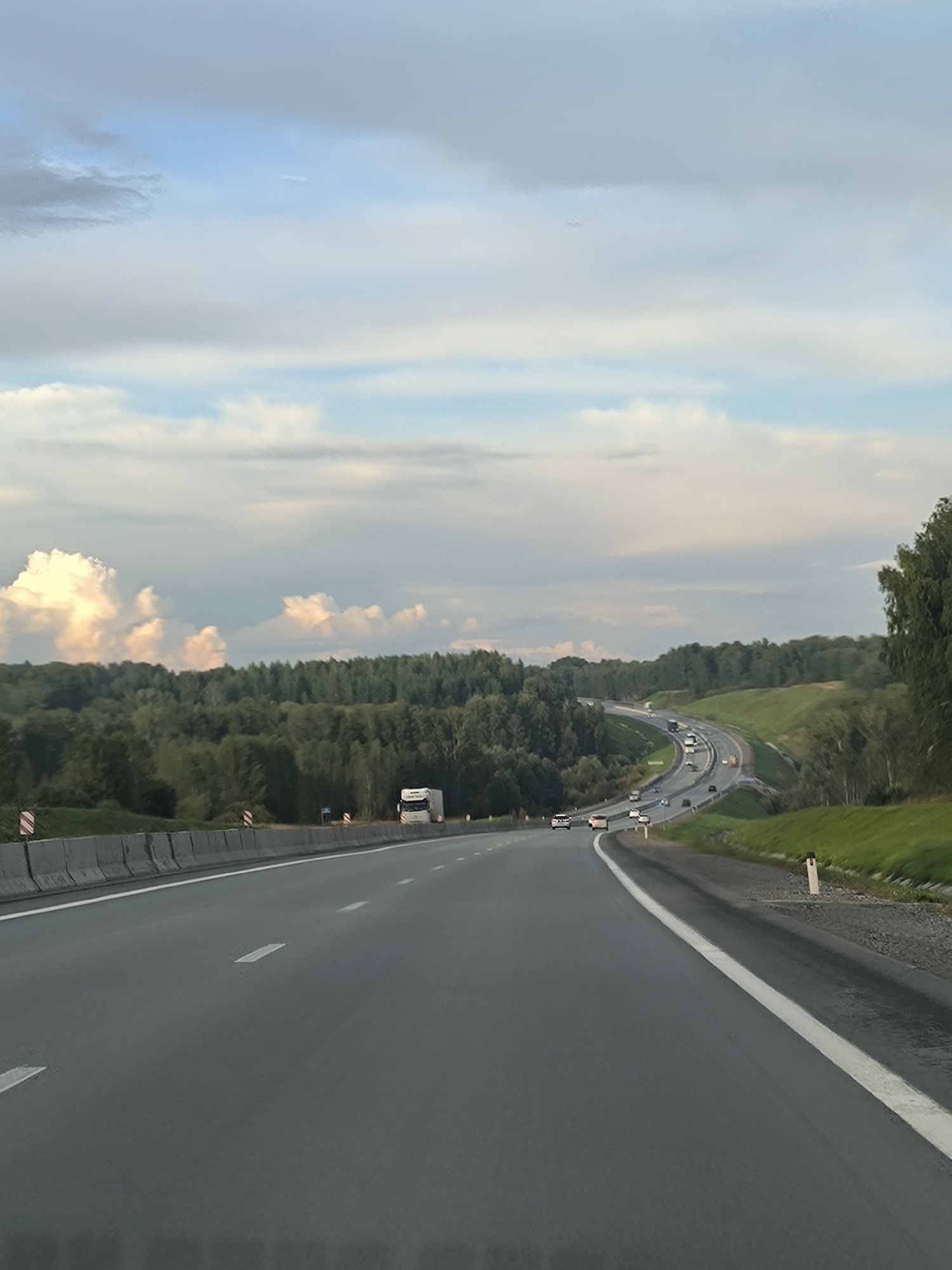 A highway stretches into the distance, curving gently through a landscape of rolling green hills and dense forests under a cloudy sky. The road is asphalt with white lane markings. Several cars and a truck are visible on the highway. The sky is a mix of blue and gray clouds, with some bright white clouds illuminated by sunlight on the left side of the frame. The scene appears to be in a rural area.