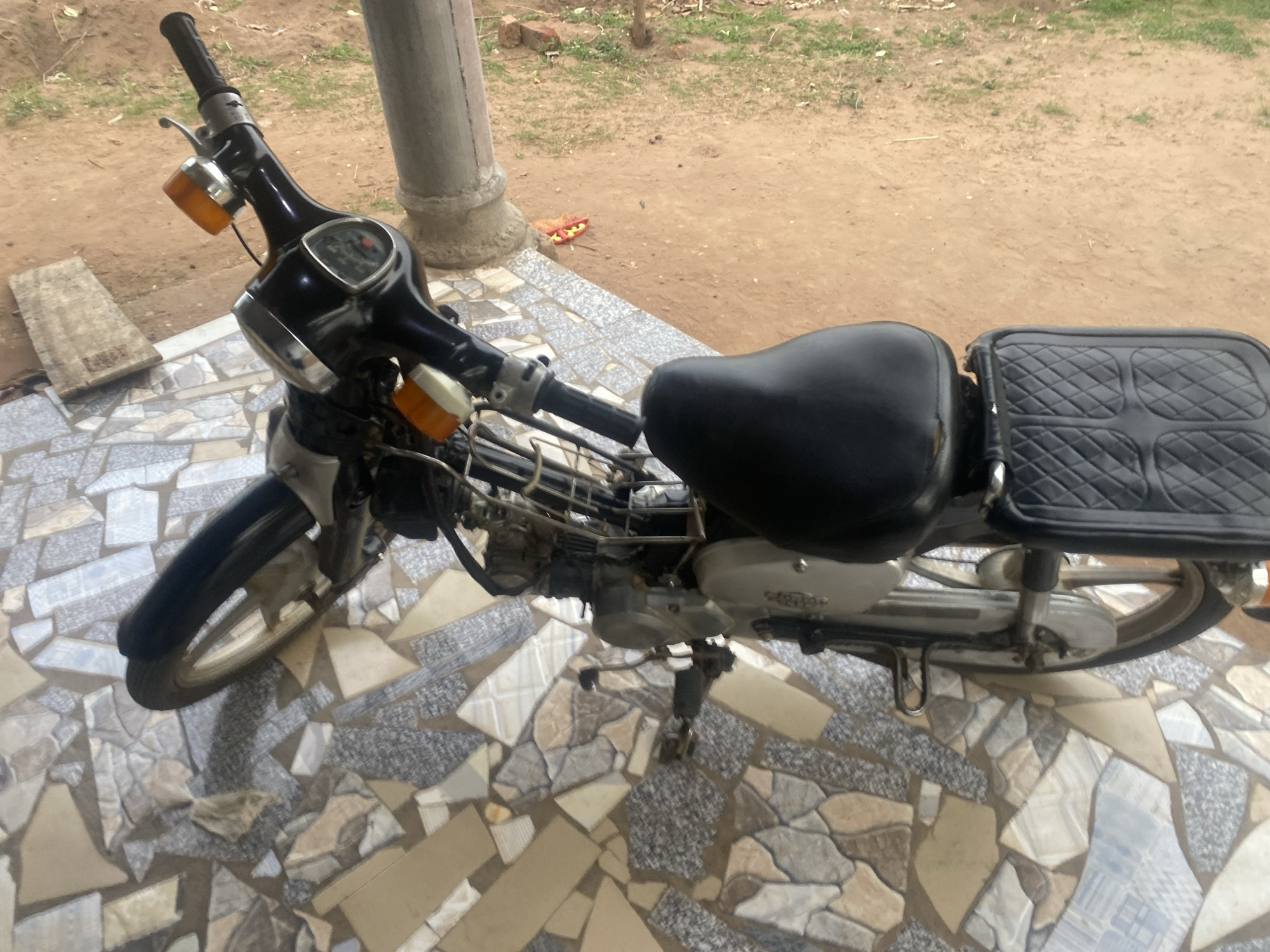 The image displays a dark-colored small motorcycle, possibly a moped or step-through scooter, parked outdoors in Makurdi, Nigeria, during daylight hours. The motorcycle is positioned on a paved area with a distinctive mosaic-like pattern, composed of irregularly shaped pieces of light-colored tiles in shades of white, grey, beige, and blue.
Key features of the motorcycle include a black single seat and a black quilted cover placed over the rear rack or fender. The handlebars are visible with a small instrument cluster and amber turn signals on either side of the front headlight housing. The front wheel and fender are partially visible, as are parts of the engine and frame towards the center. A kickstand keeps the motorcycle upright.
Beyond the tiled surface, the ground transitions to bare earth with scattered patches of green grass and small plants. A wooden plank lies on this dirt surface to the left of the motorcycle. A concrete pillar, likely part of a building structure, is partially visible on the right side of the frame, further indicating an outdoor setting. The natural light suggests it is daytime. There are no people visible, and no specific activities are taking place. No discernible text is clearly visible on the motorcycle or in the surrounding environment.