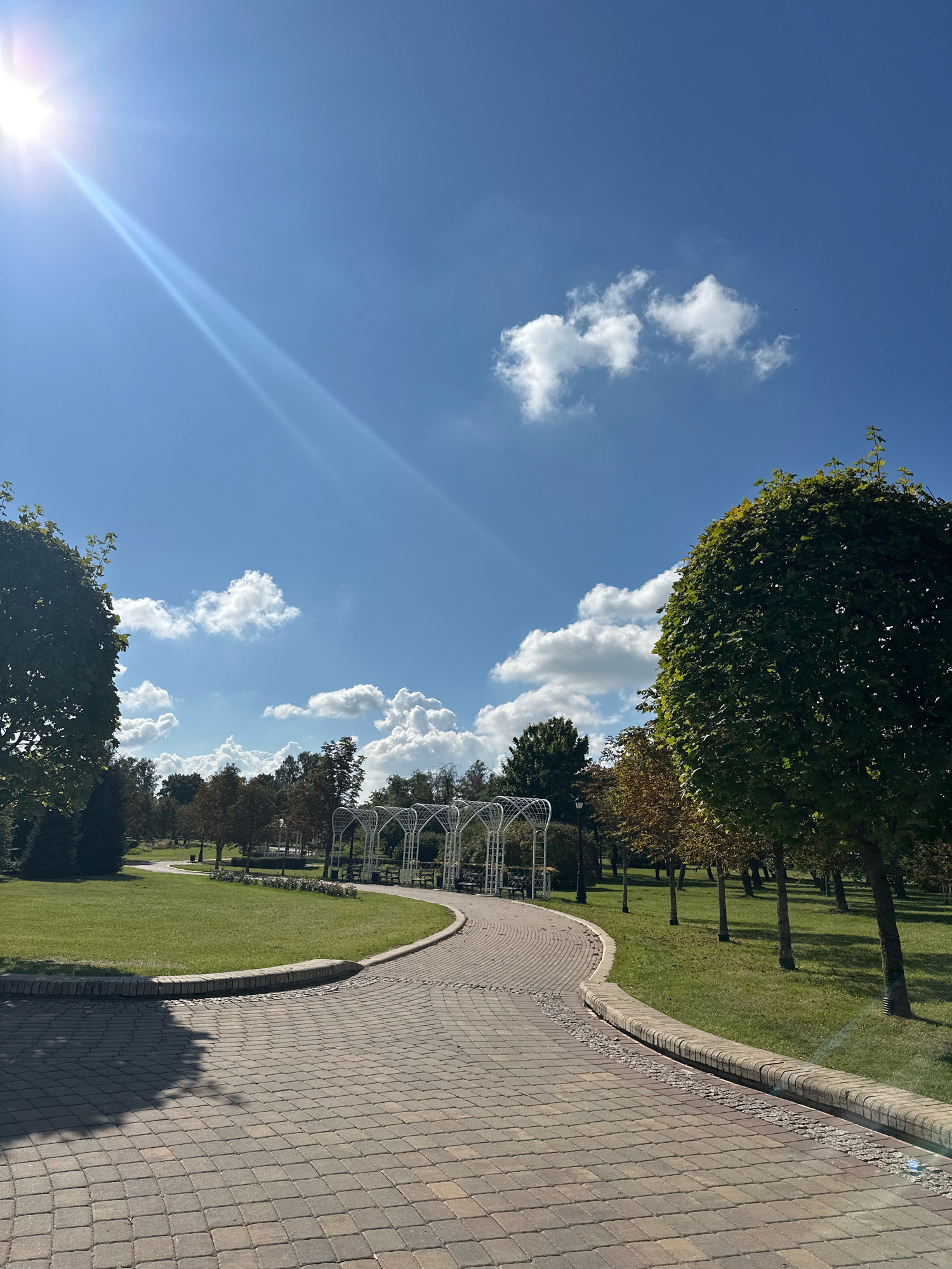 The image depicts a park scene in Minsk, Belarus. A paved pathway, constructed with rectangular brick pavers, curves through the park. The pathway is bordered by a low, curved stone wall and a strip of gravel. To the left of the pathway is a manicured lawn. In the background, several white, arched structures, possibly gazebos or pergolas, line another path. Beyond these structures, a row of trees with green and yellow foliage is visible, indicating it is likely autumn. The sky is a vibrant blue with scattered white cumulus clouds. A bright sun is positioned in the upper left corner, casting a lens flare and diagonal light rays across the scene. Large trees with dense green foliage frame the edges of the image on the left and right. The overall impression is a serene and well-maintained urban park on a sunny day.