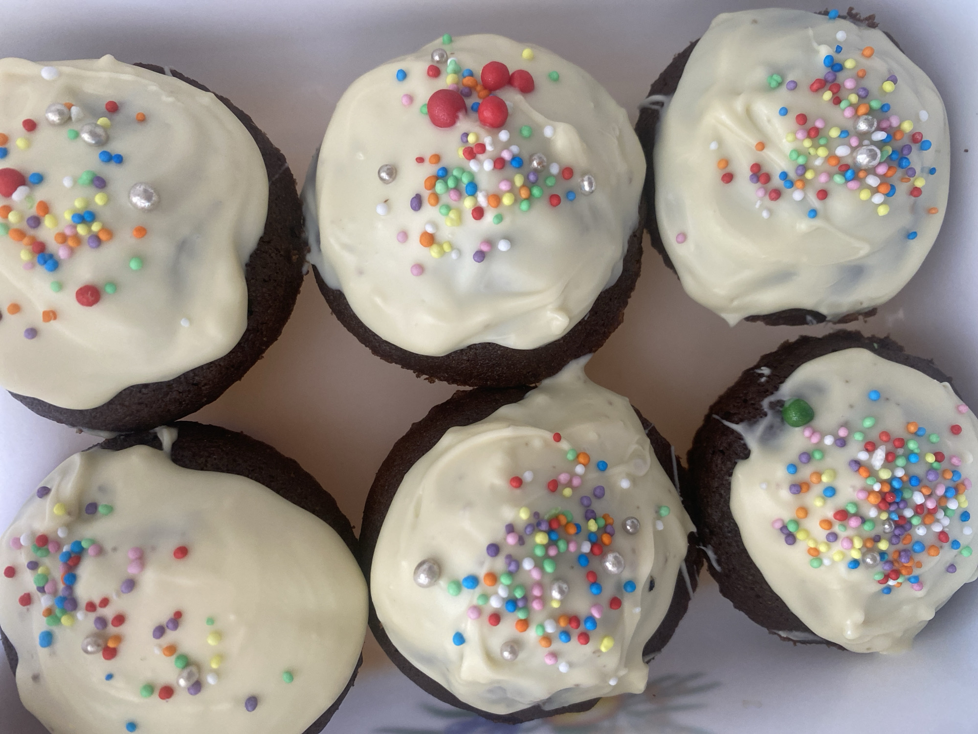 The image displays a top-down view of six chocolate cupcakes arranged in a white rectangular container. Each cupcake is topped with white frosting and adorned with a scattering of colorful sprinkles and silver edible pearls. The cupcakes appear to be homemade, with the frosting applied unevenly, creating a rustic aesthetic. There are no people or actions visible in the image. The environment is a close-up shot of the desserts, with no discernible background elements to indicate a specific location within Podgorica, Montenegro, or any other city.