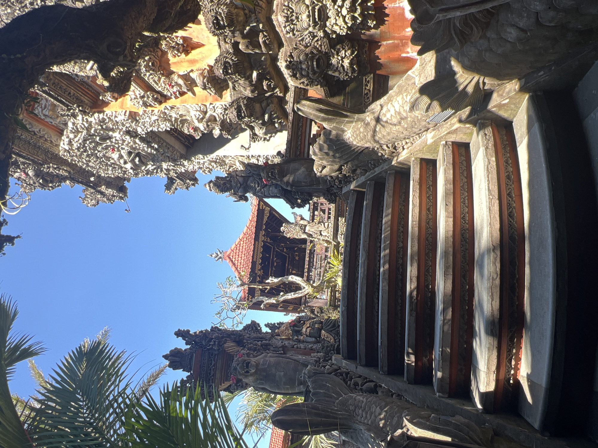 The image displays an ornate stone staircase leading to a Balinese temple complex within the Ubud District, Indonesia, under a clear, bright blue sky. In the foreground, a wide, curved stone staircase ascends, featuring gray treads and reddish-brown risers with carved decorative patterns. The stair railings are sculpted into the forms of mythical creatures, likely nagas or dragons, with visible scales and fins. Strong directional sunlight casts prominent shadows across the steps and the left railing.
At the top of the stairs, flanking the entrance to what appears to be a temple split gate, stand imposing stone guardian figures. On the left, a fierce-looking guardian statue resembling a lion or demon (Rakshasa) with an open mouth and fangs is present, adorned with a small red flower. To its right and slightly behind, a darker, seated statue is discernible. On the right side of the staircase, another tall, intricately carved guardian statue is visible, preceding a heavily decorated, reddish-orange temple wall or structure. This wall is densely covered with numerous relief sculptures of faces, figures, and elaborate patterns. A gnarled tree with bare branches stands near the center-left.
In the mid-background, a traditional Balinese pavilion (bale) with a multi-tiered, reddish-tiled roof and dark wooden columns is prominently featured. Other similar temple structures with reddish roofs are visible to the far left. Lush green palm fronds frame the upper left portion of the scene, while the upper-middle and right sections are dominated by the unblemished blue sky. No people or active interactions are depicted in the scene.