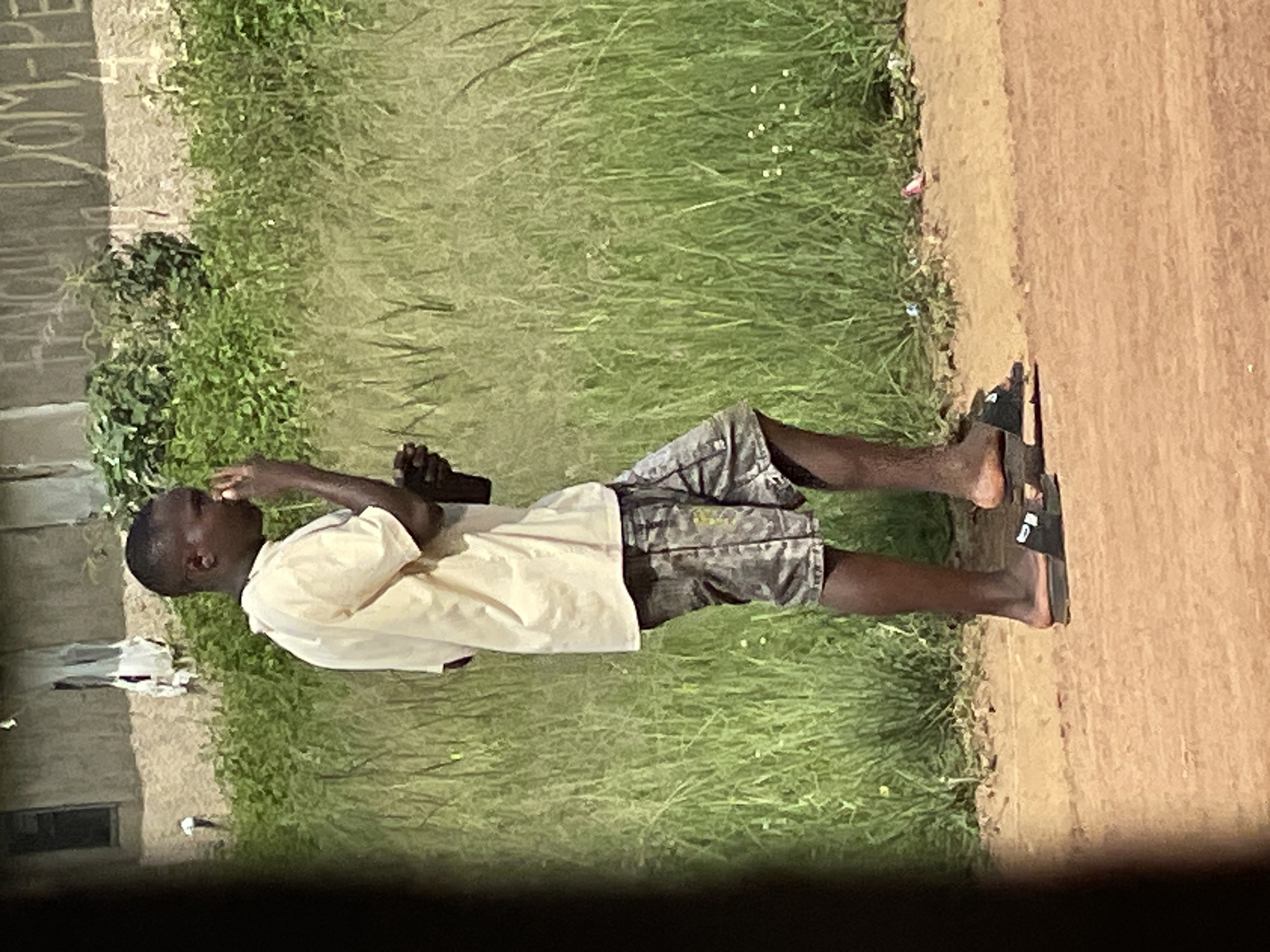 A medium shot captures a young Black male standing on a dirt road in Durumi, Nigeria. He is facing to the right, looking down and touching his nose with his right hand. He is wearing a light yellow t-shirt, grey camouflage shorts, and black flip-flops. He is holding a dark object, possibly a bottle, in his left hand. Behind him is a dense area of green grass and vegetation. In the background, a building with a textured, light-colored wall is visible, with some white cloth hanging from it. Faint writing is visible on the building's wall. The ground is a reddish-brown dirt.