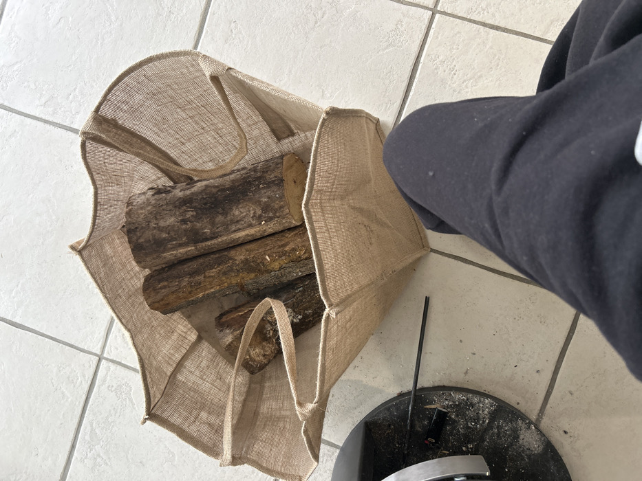 The image, captured in Veauchette, France, presents a high-angle view of an indoor scene. The lower portion of a person, identifiable by legs clad in black sweatpants, occupies the bottom right quadrant. Adjacent to the person, a beige burlap bag stands open, containing multiple cut firewood logs of varying sizes and bark textures. The bag and person are positioned on a light-colored tiled floor with a visible grid pattern. To the left, a dark, circular metallic base of an object, possibly a stove or fireplace component, is partially visible, surrounded by a light dusting of ash and debris. A thin, black, rod-like object rests horizontally on the tiled floor near the metallic base.