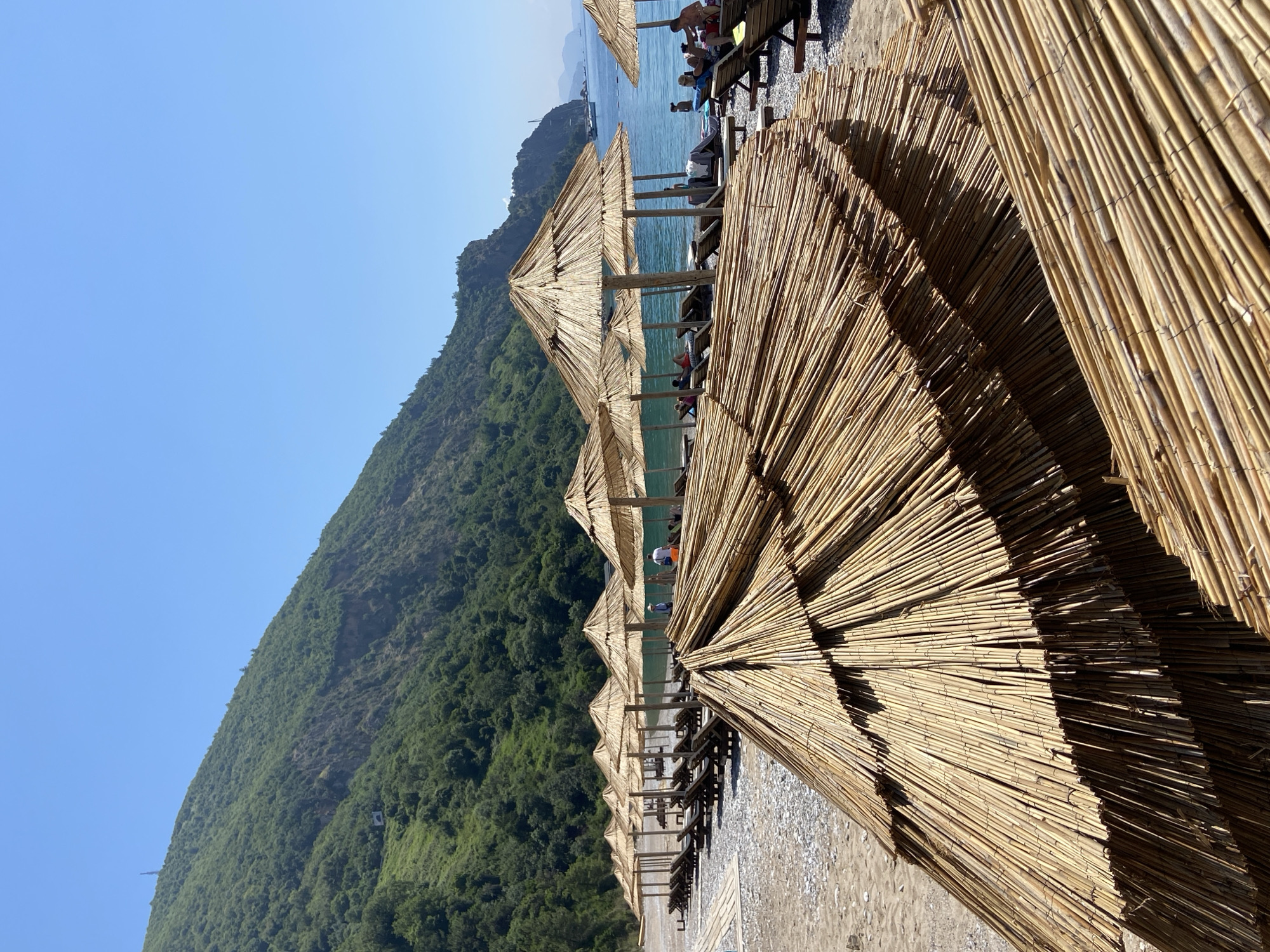 A sunny day at a beach in Prijevor, Montenegro. In the foreground, several straw umbrellas are stacked, with their bamboo poles visible. Beyond them, a row of open straw umbrellas stands over empty lounge chairs. The beach is covered in pebbles and sand. In the background, the turquoise sea is visible, with a few people sunbathing on loungers and swimming. A green, forested mountain slopes down towards the sea. The sky is clear and blue.