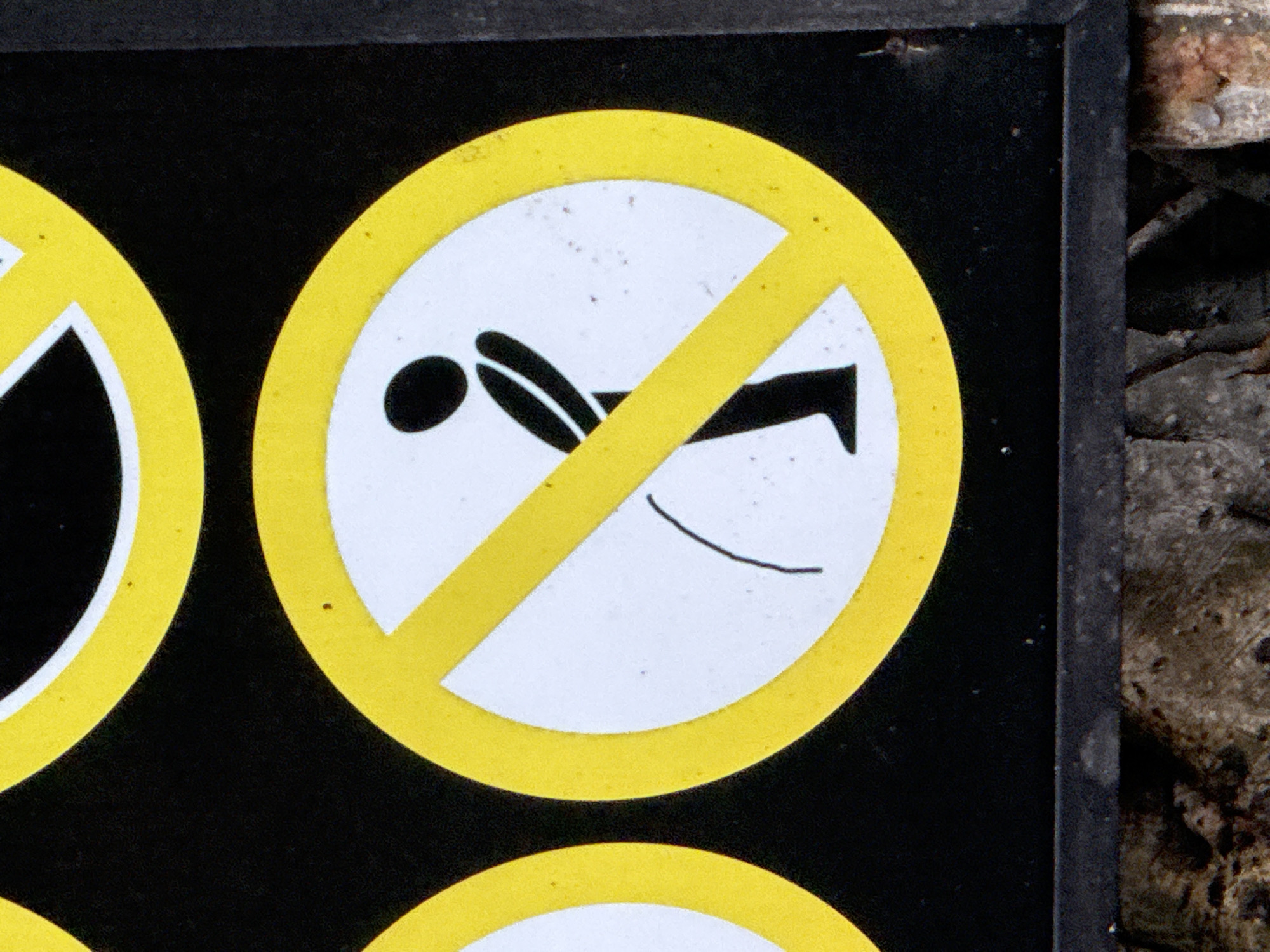 The image shows a close-up of a black sign with a yellow circle and a diagonal yellow line, indicating a prohibition. Inside the circle, a black silhouette of a person is depicted in the act of urinating, with a line representing urine. The sign appears to be part of a series of similar prohibition signs, as suggested by the edge of another yellow circle on the left side of the frame. The sign is mounted outdoors, as evidenced by the textured, earthy ground visible at the bottom of the image, which appears to be composed of dirt and possibly some fallen leaves or twigs. The overall context suggests a public space where this activity is not permitted. The image does not provide any specific information about its location in Nha Trang, Vietnam, beyond it being an outdoor public area.