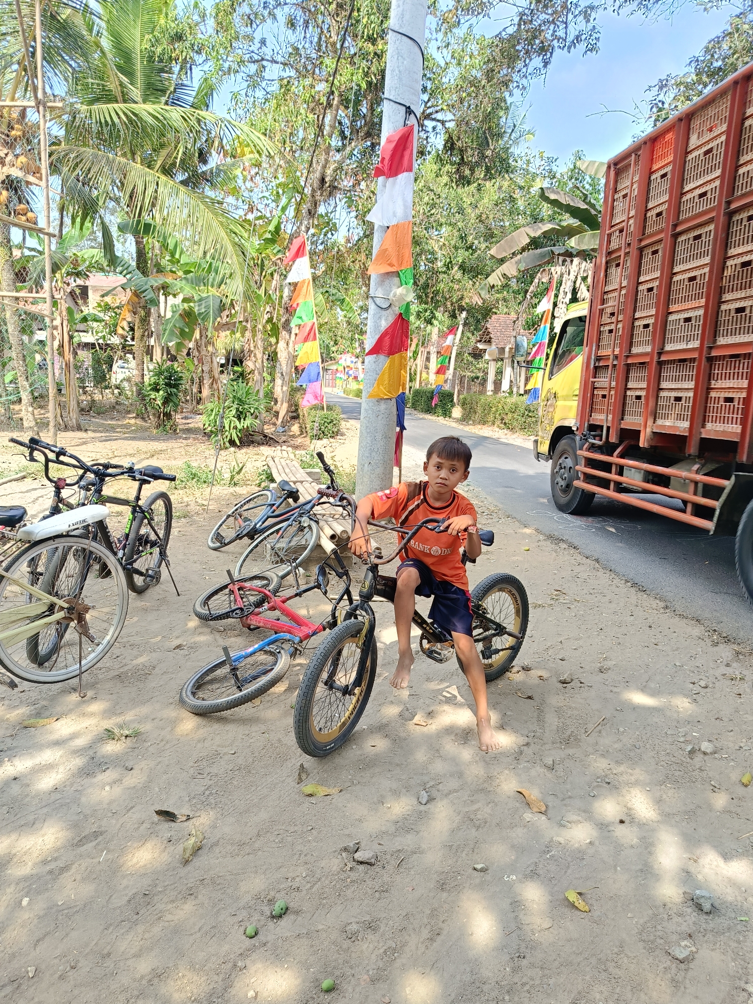 In Buayan, Indonesia, a young boy with dark hair, wearing an orange t-shirt and navy shorts, sits on a BMX bicycle. He is barefoot. The bicycle has black handlebars, a black frame, and gold rims on the wheels. Several other bicycles are parked around him on a dirt roadside. One bicycle to the left is white with a yellow rear wheel. Another is red and blue, lying on its side. A black bicycle with a white "EXOTIC" sticker is standing upright. In the background, a large yellow truck with a red cargo bed is parked on the asphalt road. The scene is illuminated by daylight and features lush green tropical vegetation, including palm trees. Decorative Indonesian flags, in red and white, are strung along a utility pole. The ground is covered in dirt, leaves, and small green fruits.