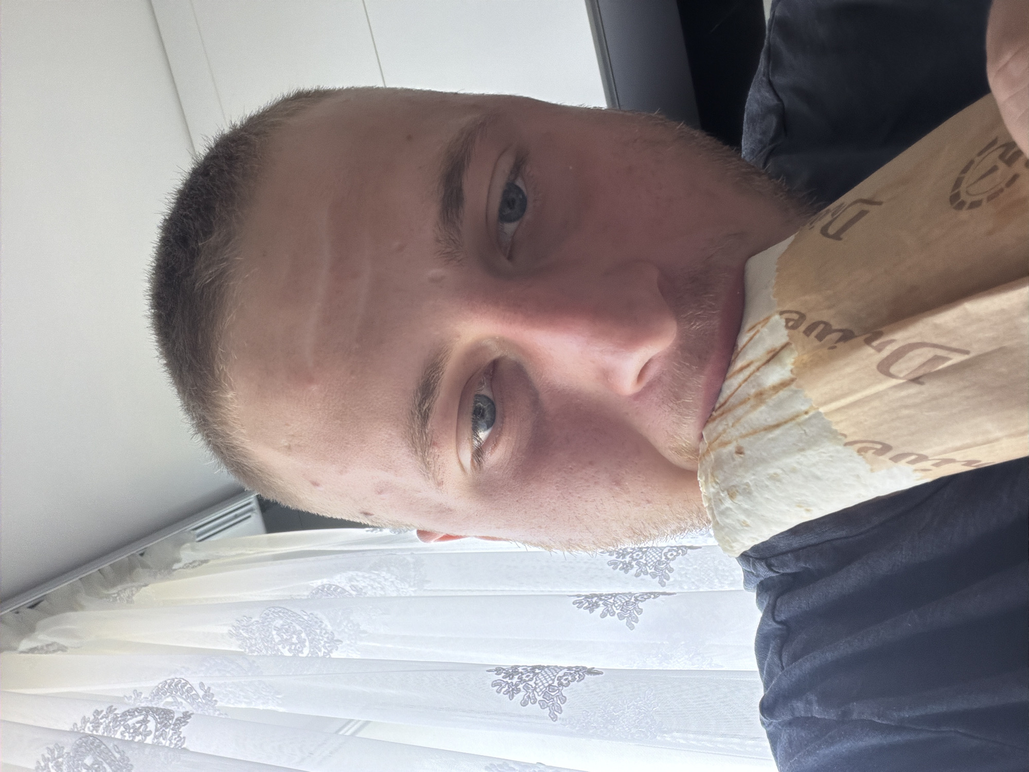 A close-up shot shows a young Caucasian male with short, light brown hair and light blue eyes. He is eating a burrito wrapped in brown paper packaging. The background features a white curtain with a decorative pattern, and a white ceiling. Part of the man's dark blue t-shirt is visible. The image appears to have been taken indoors, likely in a residential setting in the city of Strășeni, Moldova.