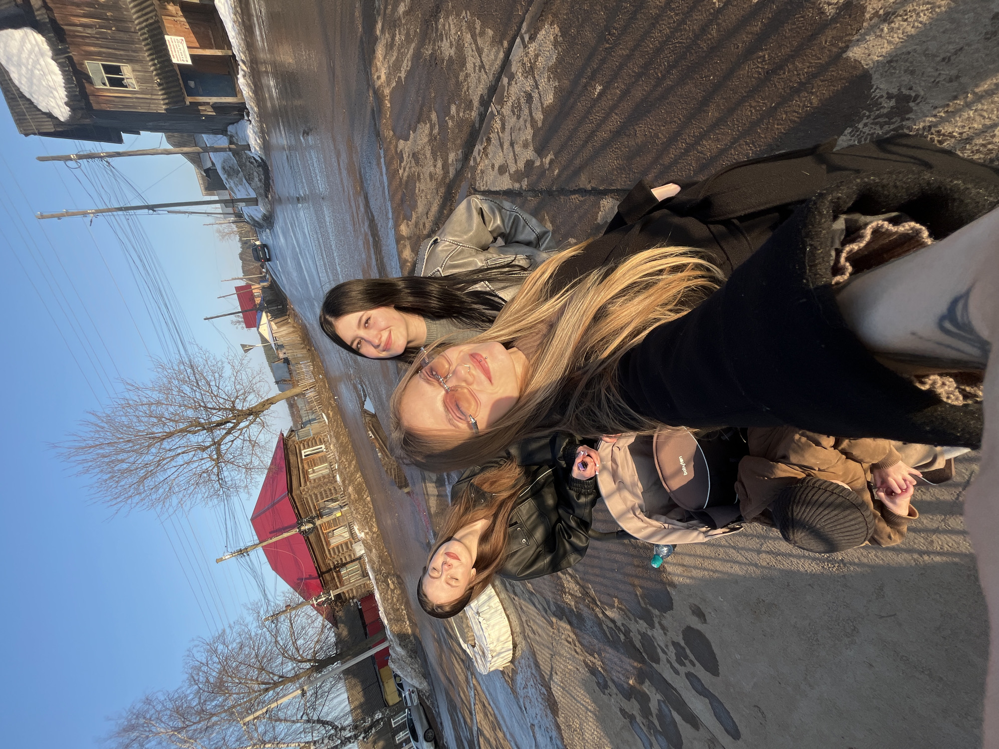 An outdoor, eye-level selfie captured in the city of Фоки, Russia, depicts three young females and an infant on a wet, partially cleared street during daylight, likely late afternoon due to long shadows.
In the foreground, a blonde female, partially visible, wears pink-tinted sunglasses and a black coat. She holds the camera, with her right arm showing a tattoo. To her left, another female with reddish-brown hair wears a black leather jacket, her eyes closed, basking in sunlight. She leans against or sits on a white tire used as a barrier. A beige baby stroller with a "babytym" label and a green plastic bottle is situated in front of her. Partially visible within the stroller is an infant wearing a brown jacket and a black knit cap, with clasped hands. Behind the blonde female, a third female with dark hair wears a grey metallic-looking jacket and smiles towards the camera.
The immediate ground is wet asphalt or packed dirt, showing puddles and reflections, consistent with melting snow. Long, distinct shadows are cast across the ground from the subjects and unseen objects. In the midground, the street ascends slightly. Patches of dirty snow and ice are visible along the road edges and near structures.
The background features several wooden buildings under a clear blue sky. To the left, a log cabin-style house with a bright red metal roof is prominent. Utility poles with multiple electrical wires run overhead and along the street. Further down the street on the right, an older, darker two-story wooden building with a corrugated roof and a snowdrift on its roof is visible. A dark sedan is parked on the street in the distance. Bare deciduous trees, possibly birches, are scattered throughout the scene, suggesting late winter or early spring conditions.
