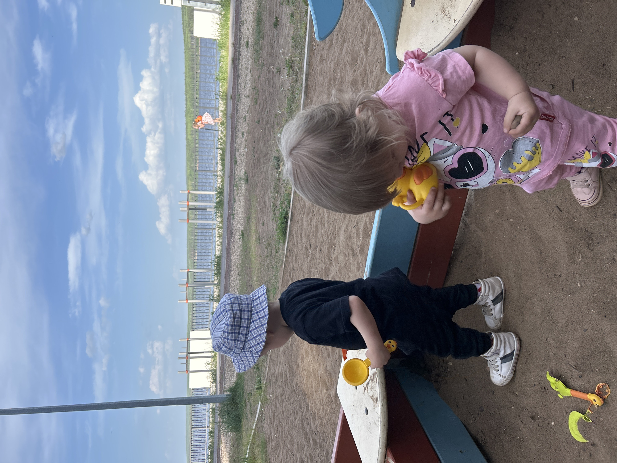 Two young children play in a sandbox at an outdoor playground in Chaikovsky, Russia. The child on the right, with blonde hair, wears a pink shirt and light pink sneakers and holds a yellow toy. The child on the left, wearing a blue checkered sun hat, black shirt, black pants, and white sneakers, is seen holding a yellow scoop. The playground has colorful borders, and the backdrop reveals a partially visible fence and a clear blue sky with scattered clouds.