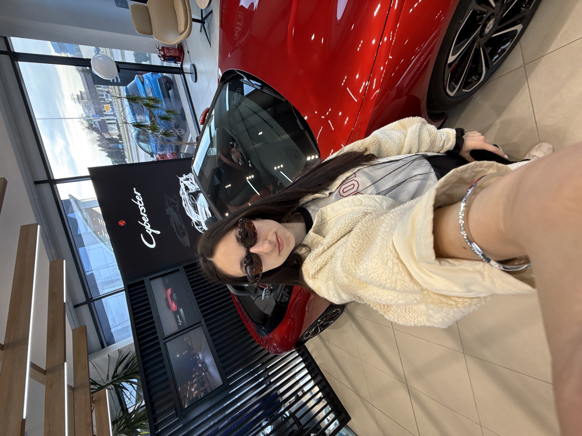 A young Caucasian female with long, dark brown hair and dark oval sunglasses is depicted taking a selfie inside a car showroom. She wears a cream-colored fleece jacket over a light shirt with a red graphic. A silver-toned bracelet is visible on her right wrist, while her left arm extends forward, holding a mobile device for the photograph. Behind her, a glossy red MG Cyberster convertible sports car is displayed on a light-tiled floor, featuring black multi-spoke wheels and a brown/tan interior. A large black vertical display board immediately behind the car showcases MG and Cyberster branding, alongside car diagrams. Two smaller screens to the left of the main display show additional car images. The showroom ceiling incorporates linear LED lights within wooden panels. Large windows reveal an exterior street scene with parked vehicles, including a blue hatchback bearing a Bulgarian license plate "CB 7706 CB." Across the street, a building with a green roof and a sign partially displaying Cyrillic text are visible. The license plate and Cyrillic script confirm the location as a car dealership in Sofia, Bulgaria. Within the showroom, to the right, a beige armchair, a circular side table, a white spherical floor lamp, and a red fire extinguisher are present.