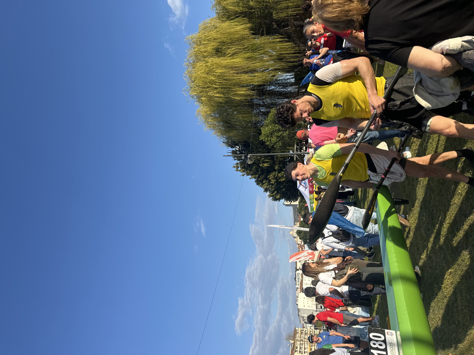 The image depicts an outdoor sporting event or gathering in Mirandela, Portugal, under a clear blue sky with sparse clouds. The scene is primarily composed of a grassy area populated by numerous individuals.
In the foreground, two young adult males stand prominently, both wearing yellow sleeveless athletic tops and dark shorts. The man on the left, wearing a black baseball cap, holds a black and yellow canoe paddle and looks forward with a slight smile. To his right, the second man, with dark curly hair, holds a similar paddle and looks down, seemingly examining his equipment. A bright green canoe or kayak hull rests on the grass to the left of these individuals.
The midground features a dispersed crowd of people in various casual attire, standing and interacting. Partial views of white and red flags or banners are visible, along with parts of white tents, suggesting event infrastructure. A white sign with the number "180" and the text "JOGOS" is partially visible on the far left.
In the background, a residential area with multi-story buildings is situated on a gentle incline. A distinctive light-colored building with a dome or spire is visible on the distant horizon. A dense line of trees extends horizontally across the middle ground, with a large weeping willow tree, characterized by its bright yellow-green drooping foliage, dominating the right side of the frame. The overall lighting suggests a sunny daytime setting.